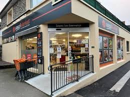 Extérieur d'un magasin de proximité avec une façade vitrée, une bannière indiquant 'Soyez Les Bienvenus', et une personne poussant une chariot dans la zone piétonne.