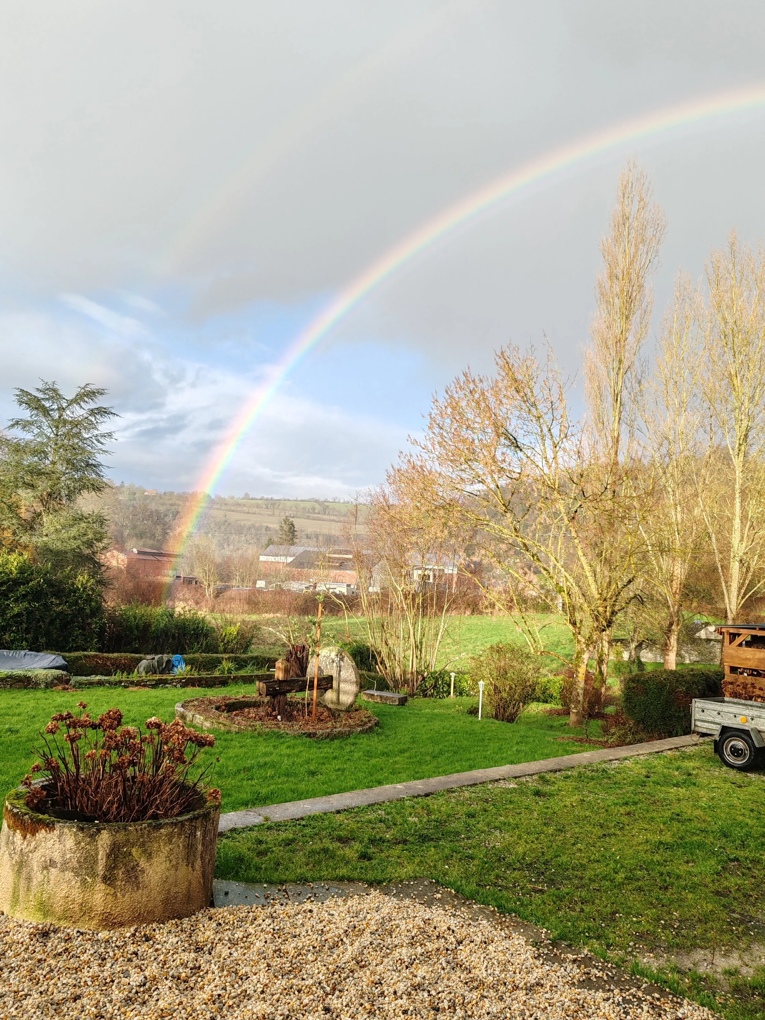 Paysage de jardin avec un arc-en-ciel double dans le ciel, arbres sans feuilles, pelouse verte, et mobil-homes en arrière-plan.