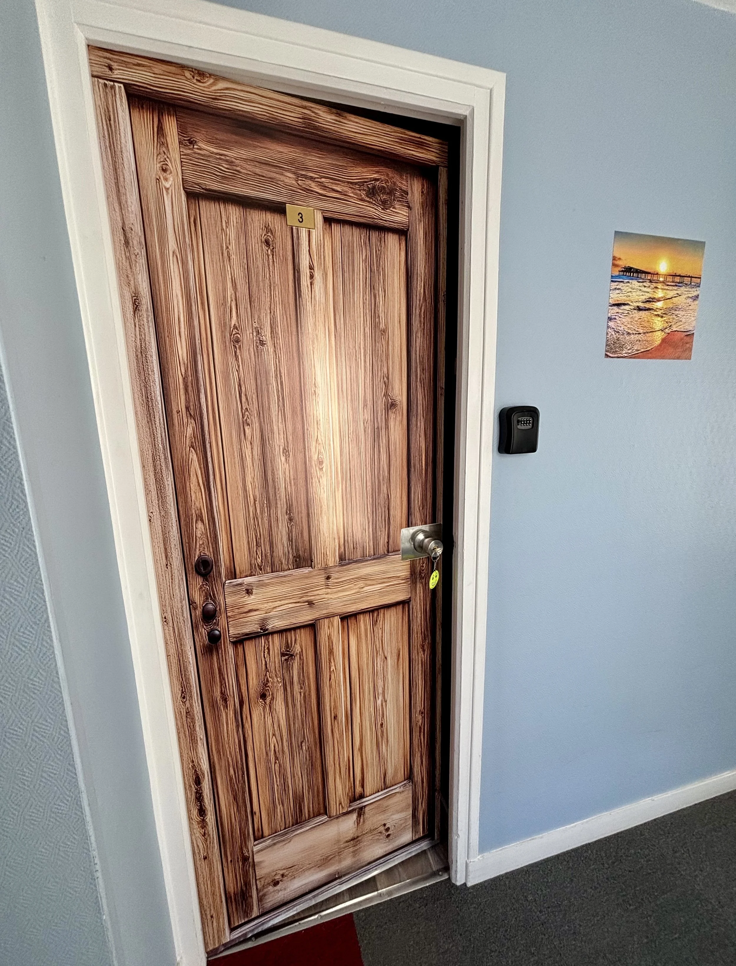Une porte en bois avec un petit panneau portant le numéro 3, une poignée de porte en métal, et un clavier numérique à proximité on la mur en bleu clair. Il y a aussi une photo de coucher de soleil sur la plage collée au mur à droite.