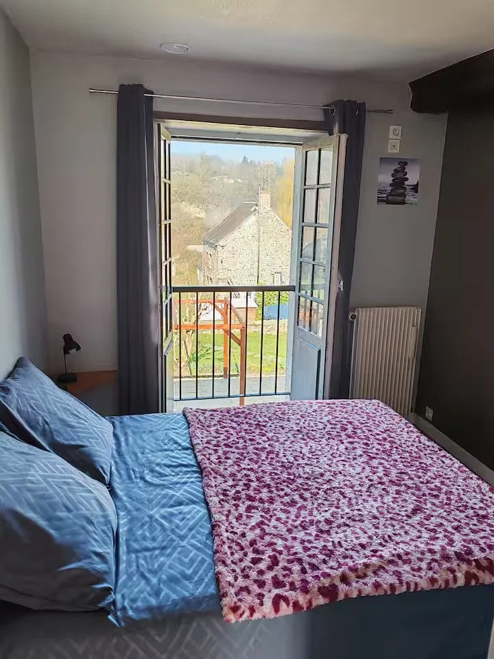 Chambre avec un lit en face d'une grande fenêtre ouvrant sur un jardin, avec des draps bleus et une couverture rose à motifs. Décor simple avec peinture en noir et blanc sur le mur, radiateur et une petite lampe de lecture.