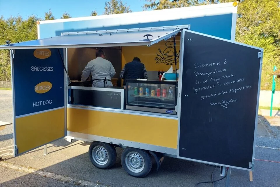 Un food truck en plein air avec deux chefs au travail, une porte ouverte avec menu affiché, et un panneau noir avec un message écrit en français