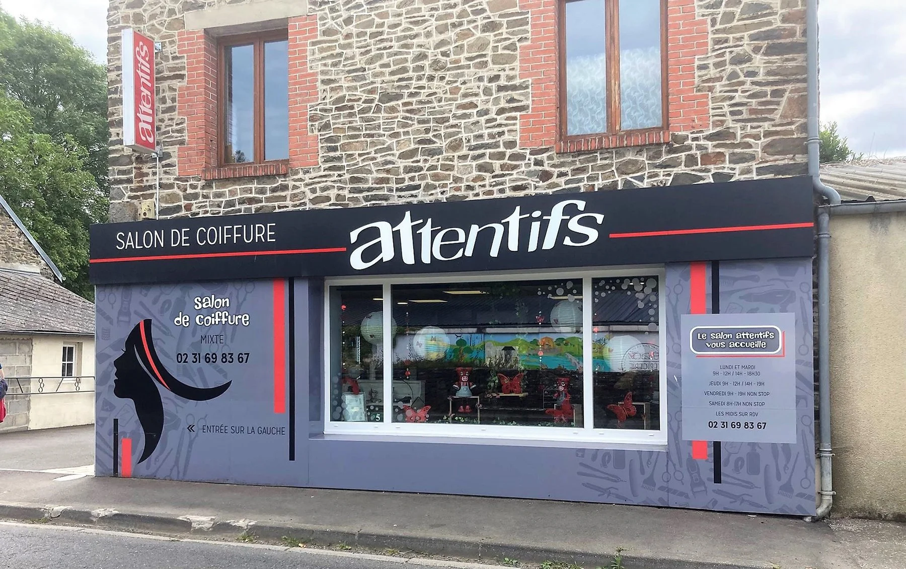 Vitrine et façade d'un salon de coiffure nommé "Alentifs" situé dans un bâtiment en pierre avec une enseigne noire et rouge. La vitrine montre des décorations colorées à l'intérieur et des informations sur les horaires d'ouverture et le contact à droite.