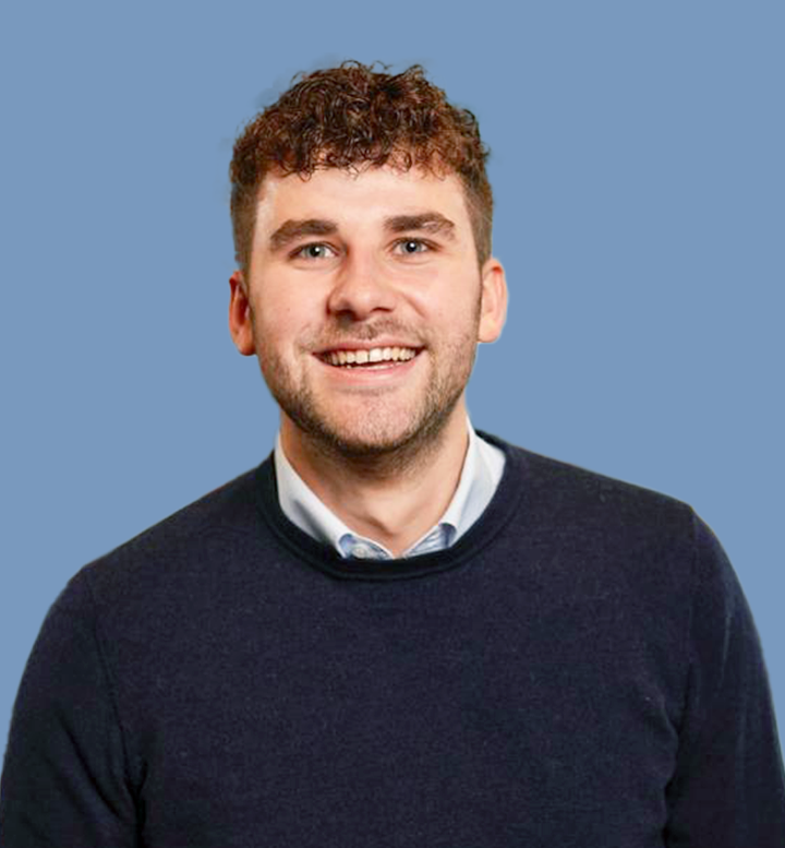 Portret van een jonge man met bruin, krullend haar, glimlach, blauwe achtergrond, donker trui en witte blouse.