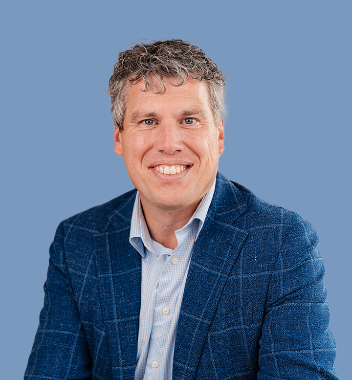 Portret van een lachende man met grijs, krullend haar, gekleed in een blauwe blazer en een lichtblauw overhemd, tegen een effen blauwe achtergrond.