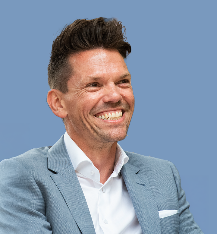 Man in een lichtblauw pak en wit overhemd lachend, op een blauwe achtergrond.