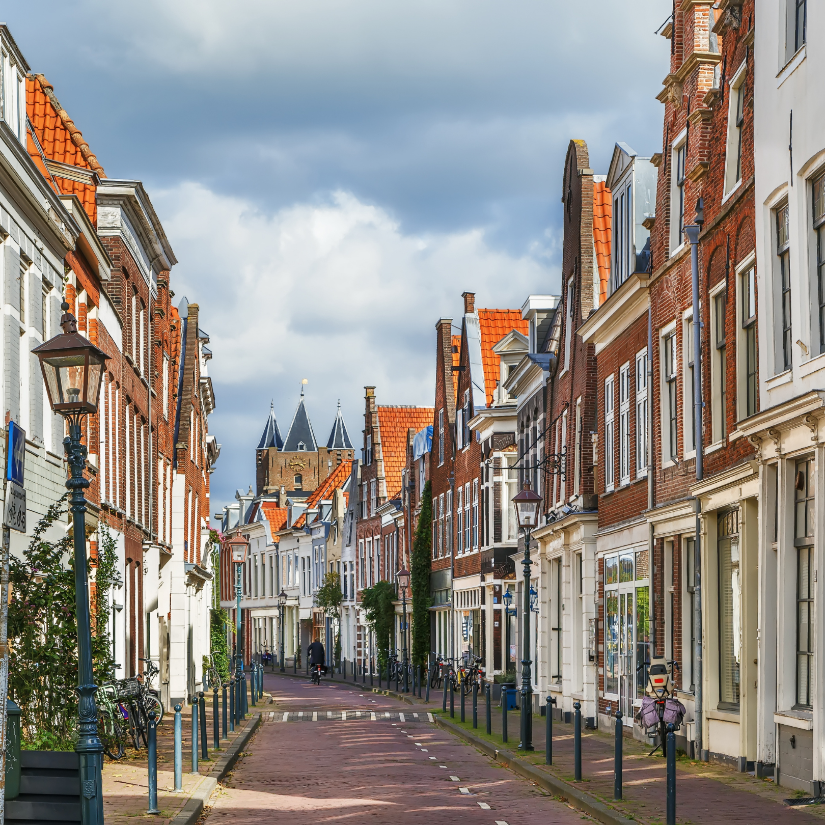 Een straat met historische gebouwen met rode daken, fietsen langs de stoep en lantaarnpalen, onder een bewolkte blauwe lucht, in een Europese stad.