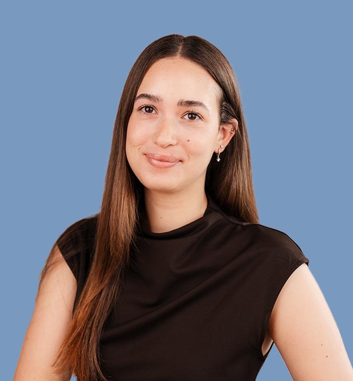 Portret van een jonge vrouw met lange bruin haar, die glimlacht en een zwarte top draagt tegen een blauwe achtergrond.