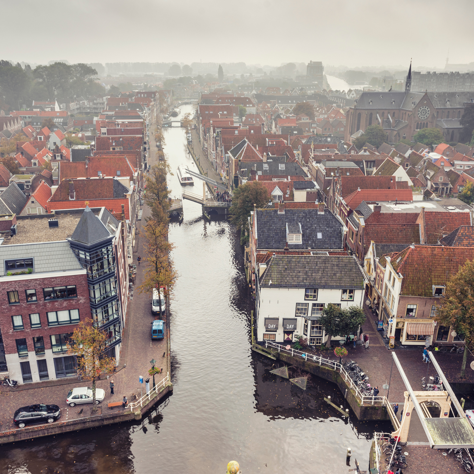 Luchtfoto van een stad met een kanaal, bruggen, huizen met rode en zwarte daken, een grote kerk en bomen in de herfst, mistige achtergrond.