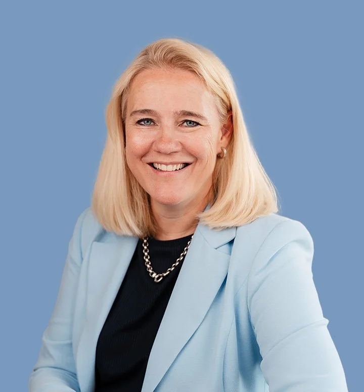 Portret van een lachende vrouw met blonde haar, gekleed in een lichtblauw blazer en een zwarte top, tegen een effen blauwe achtergrond.