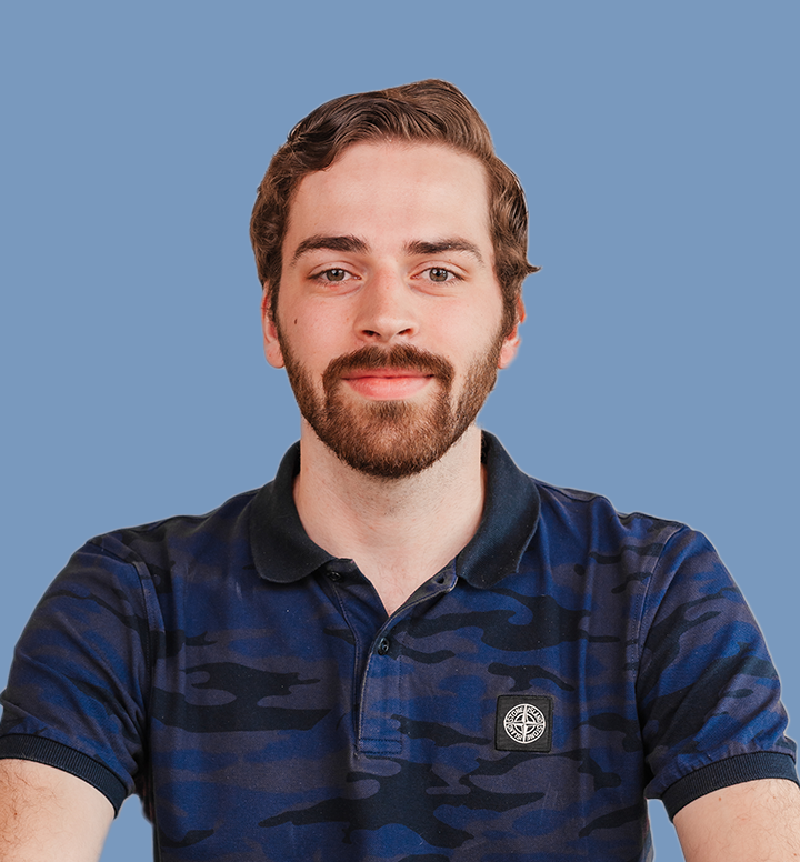 Portret van een jonge man met een baard en kort, golvend bruin haar, glimlachend, gedragen in een donkerblauwe camouflagedhirts met een logo, op een blauwe achtergrond.