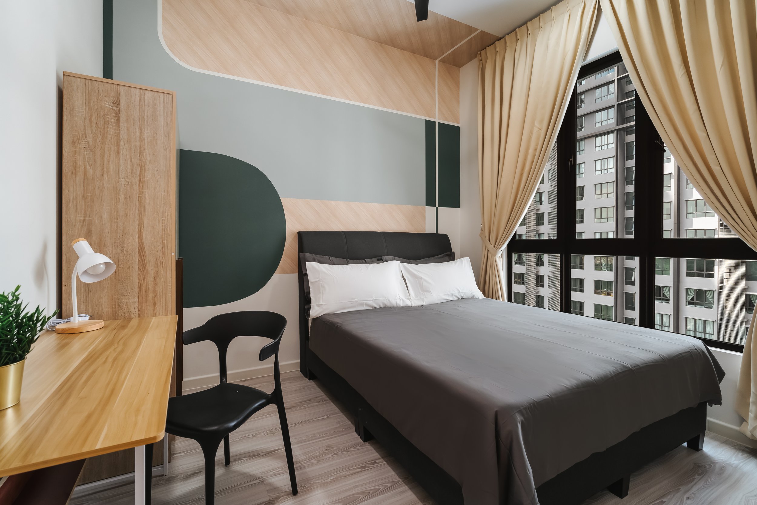 Modern bedroom with large window, beige curtains, bed with gray bedding, wooden desk with black chair, and geometric accent wall