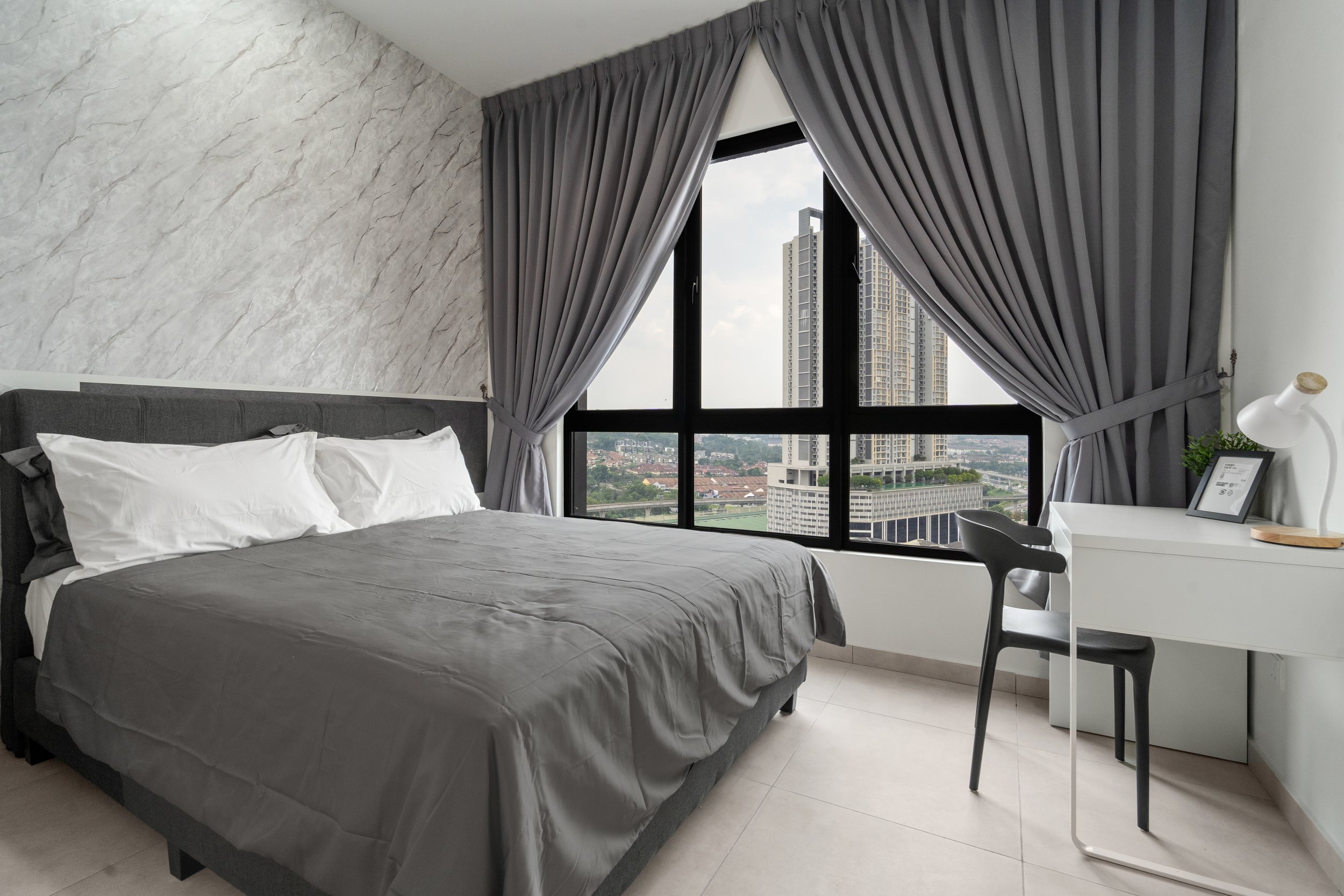 A modern bedroom with a large bed, gray bedding, and pillows, a marble-textured accent wall, black framed window with cityscape view, gray curtains, a white desk with a black chair, and a table lamp with a picture frame and green plant.
