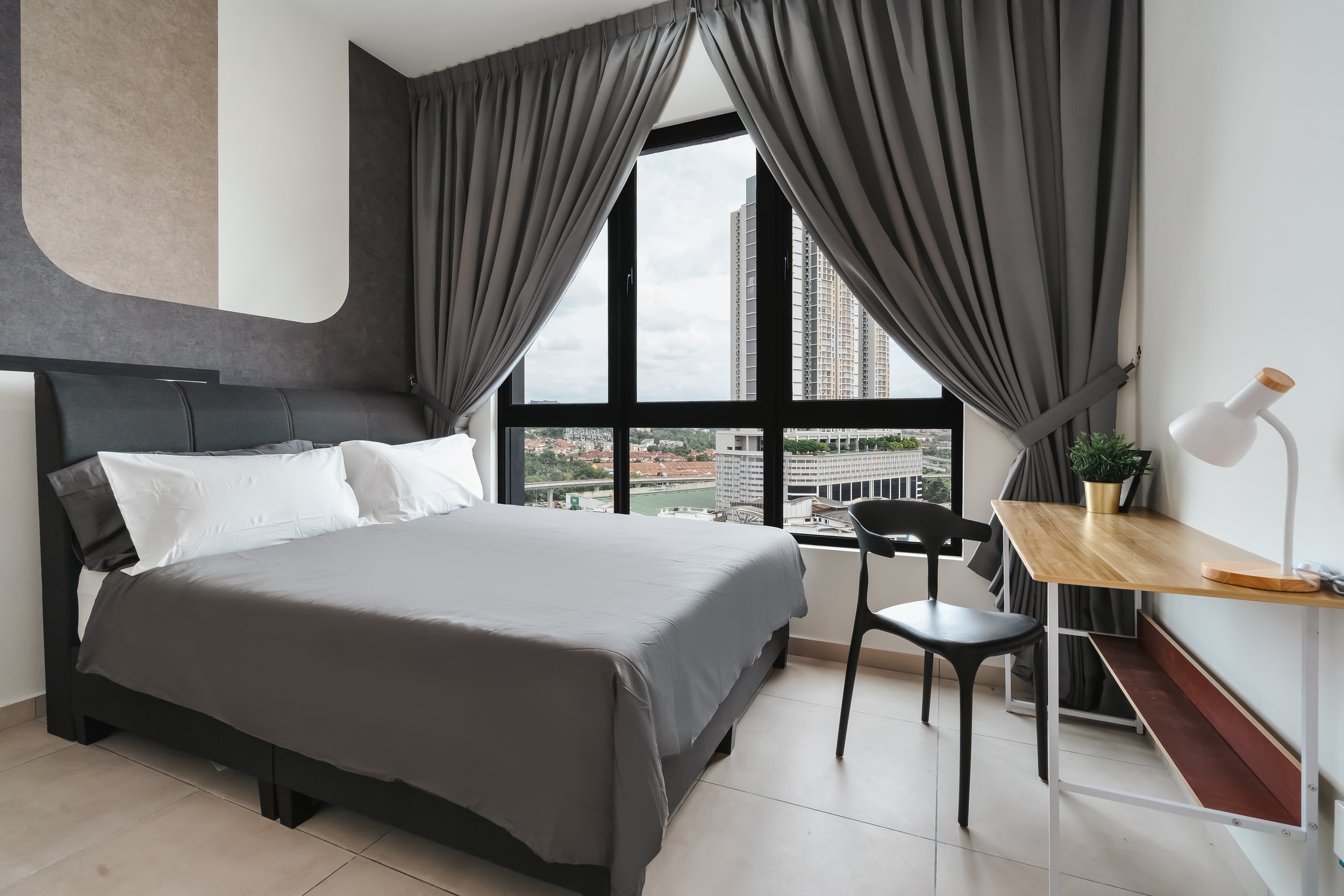 Modern hotel room with a bed, a window with city view, gray curtains, a small wooden desk with a potted plant and a white desk lamp, and black chairs.