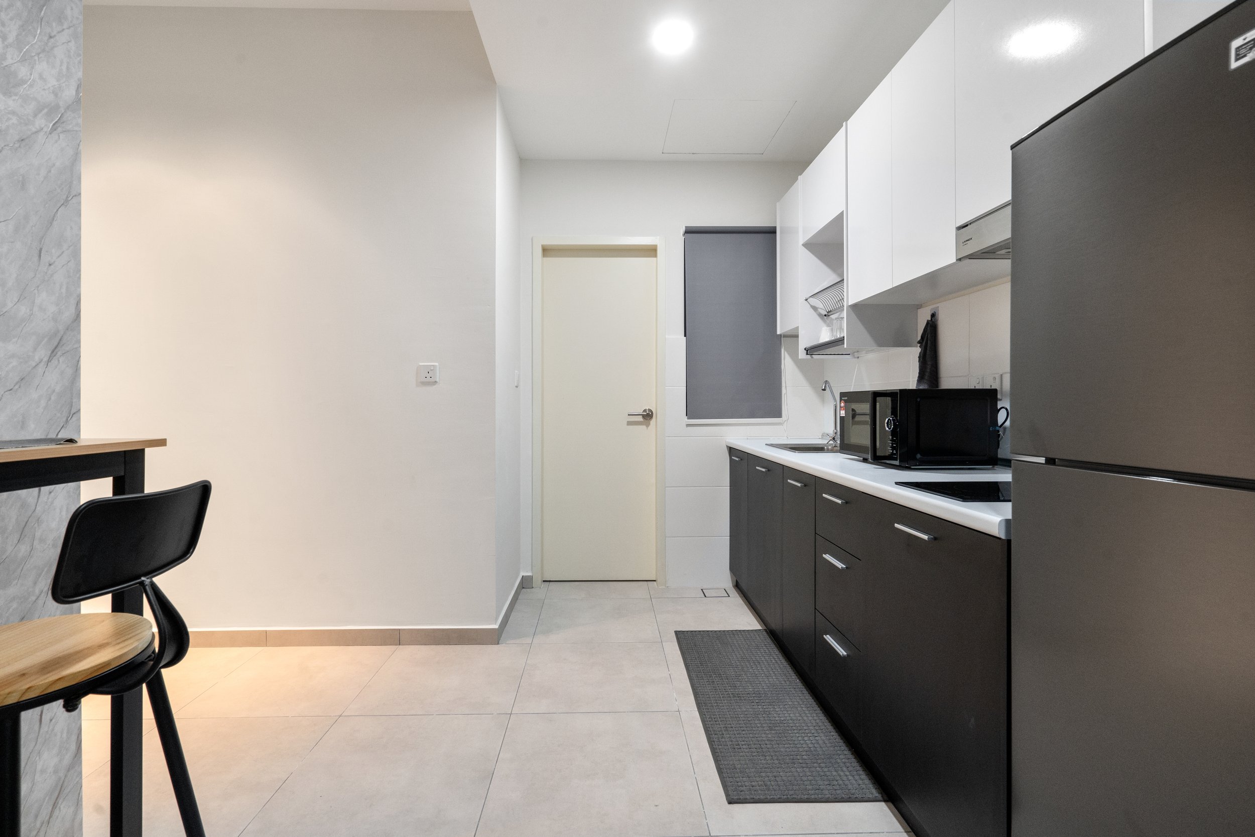 Modern kitchen with black cabinets, white backsplash, microwave, refrigerator, and a small gray rug on beige tile floor.