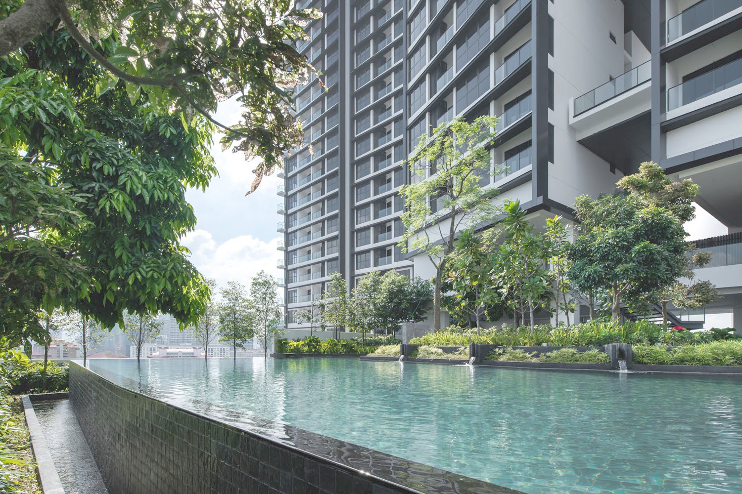 A modern high-rise condominium building with glass balconies, surrounded by green trees and a landscaped garden, with a curved swimming pool in the foreground.