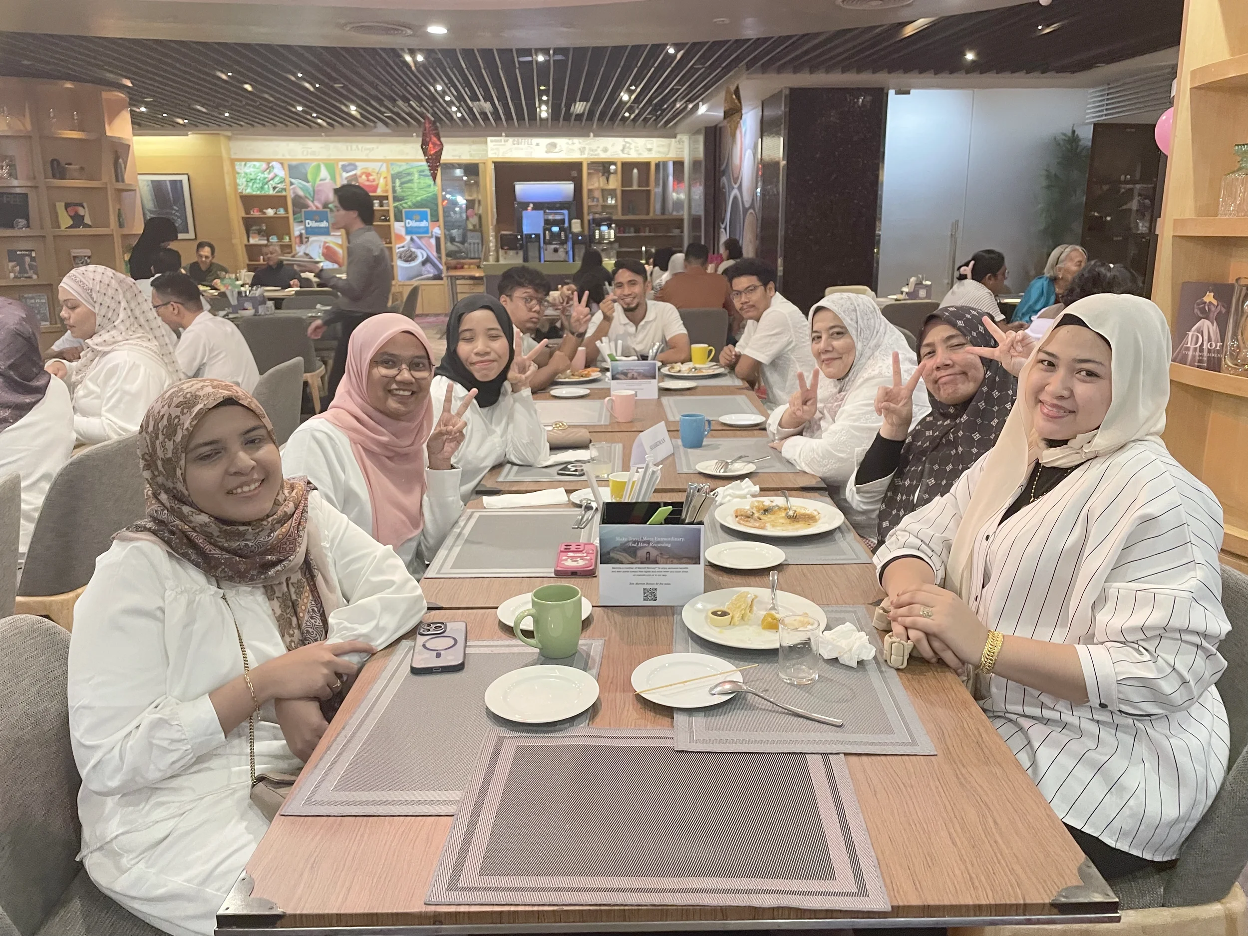 A group of people sitting at a restaurant table, smiling and making peace signs. The table has plates with food, cups, and utensils. In the background, there are other diners, a waiter, and a restaurant interior with shelves and food posters.