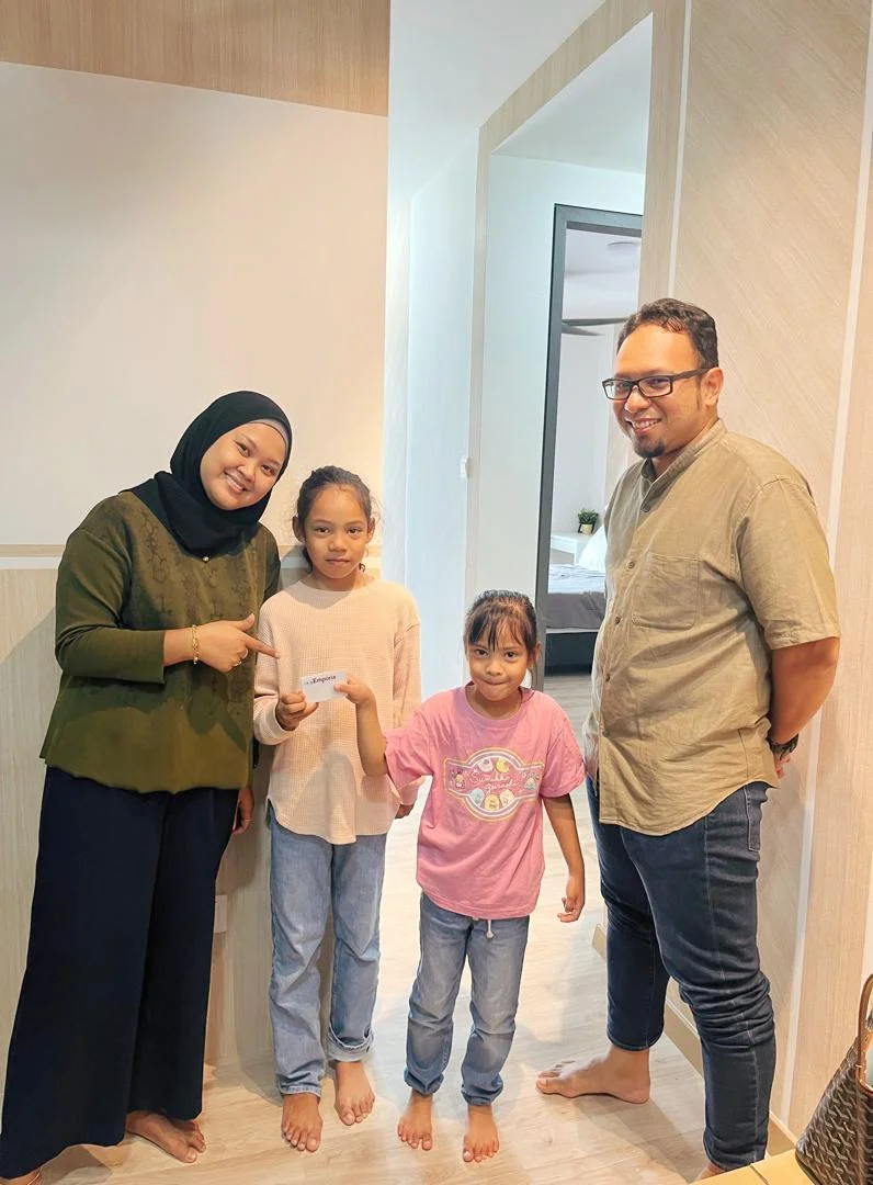 Group of four people, including two women and two girls, standing in a home interior. One girl is holding a card. All are barefoot, smiling or looking at the camera.