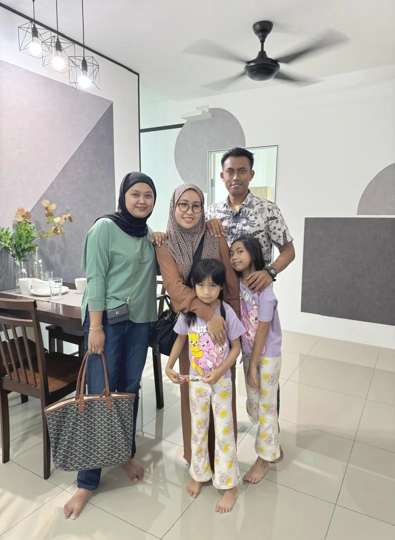 A family of five posing together indoors, including two adult women, one adult man, and two young girls, with a dining area and modern decor in the background.