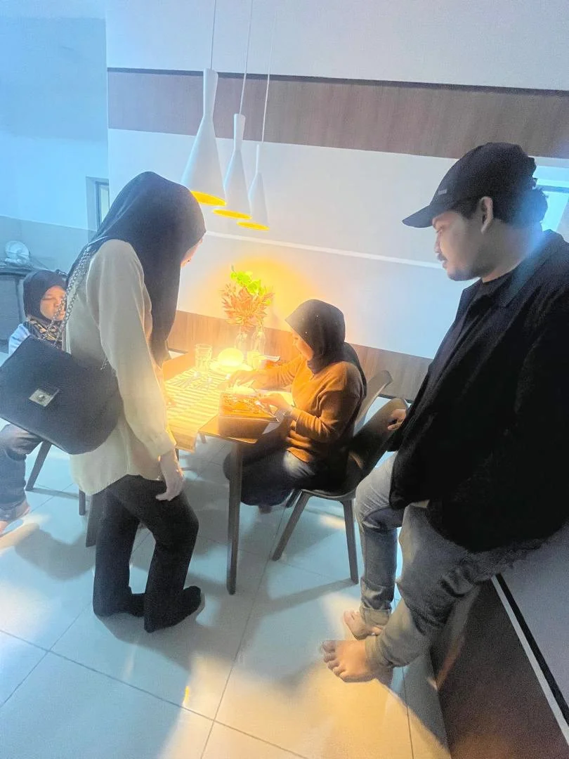 People in a restaurant or cafe, with some standing and others sitting at a table, illuminated by warm lighting.