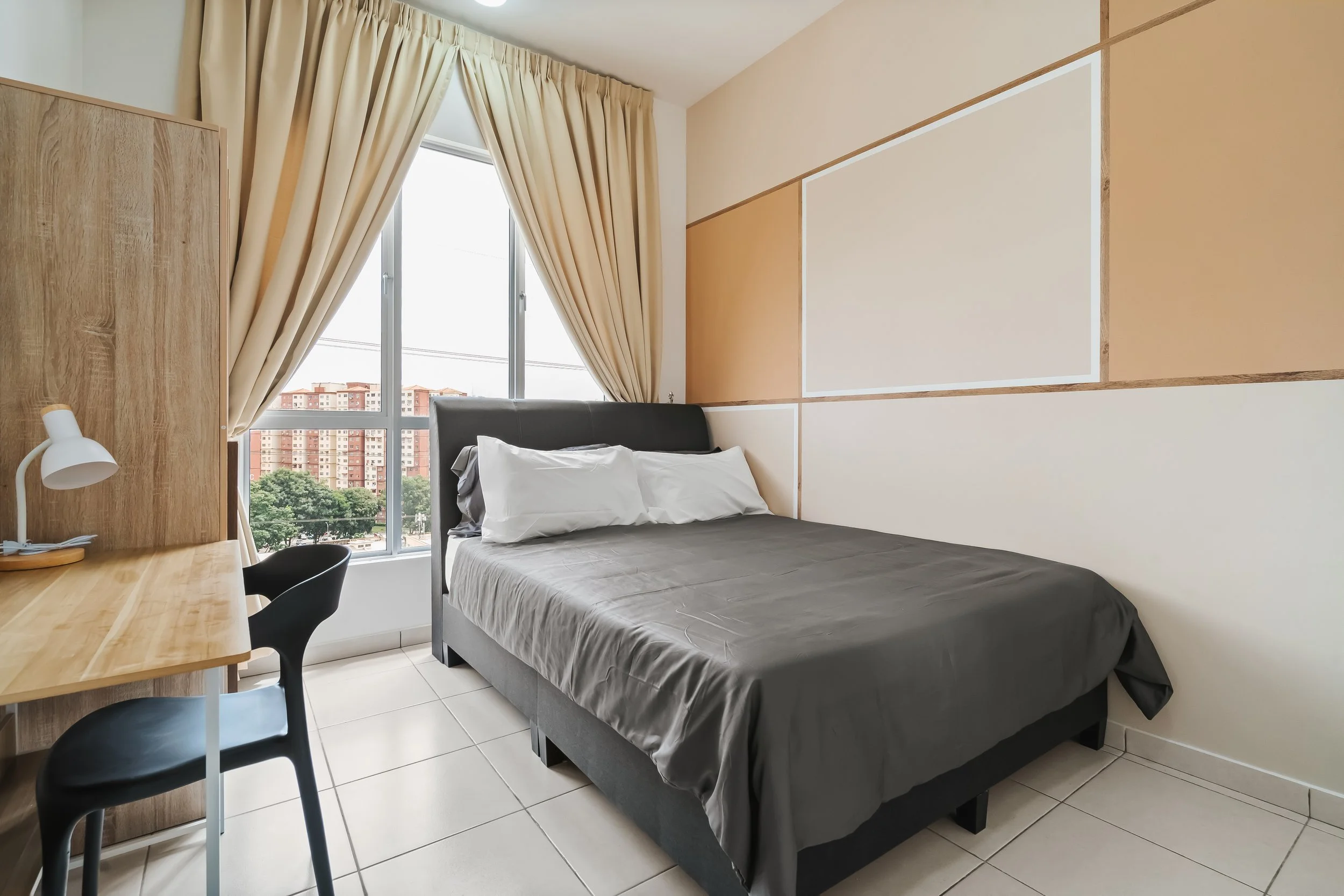 A minimalist bedroom with a large window, beige and white paneled walls, a gray bed with white pillows, a wooden desk with a white desk lamp, and black chairs. Outside the window, there are residential buildings and trees.
