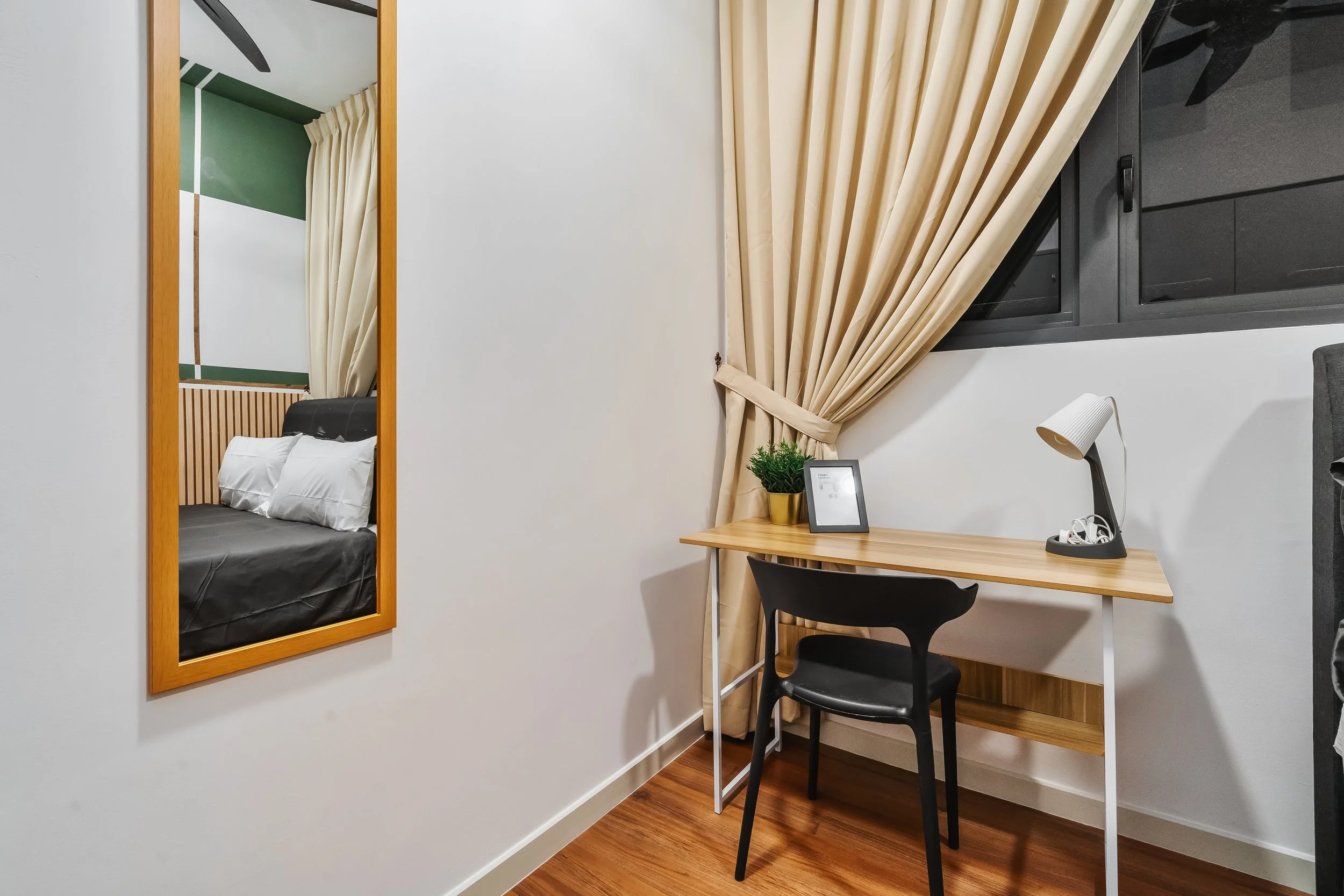 A cozy corner in a modern apartment with a small wooden desk, black chair, white lamp, potted plant, and a framed picture. There is a window with beige curtains and a mirror reflecting a bedroom with a bed, white pillows, and a headboard.