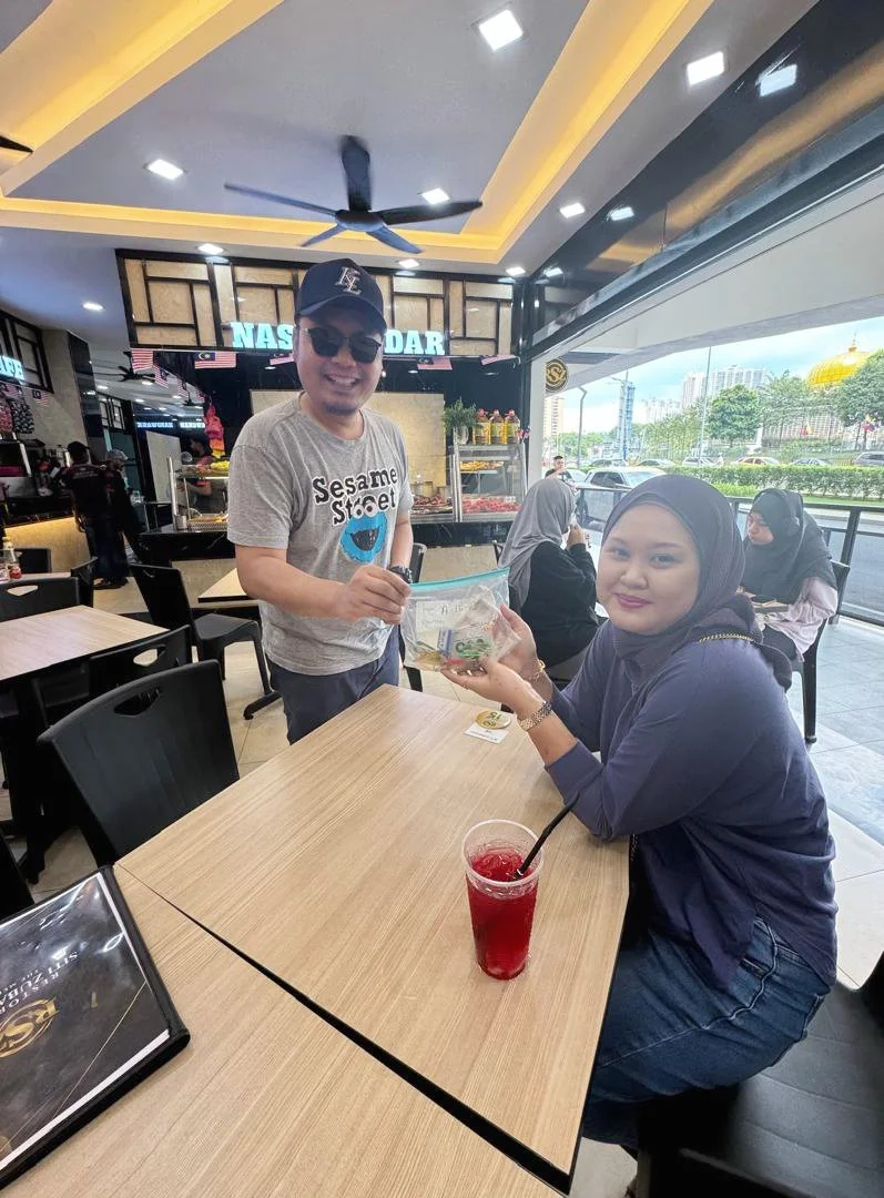 A man in a gray T-shirt with a Sesame Street mask logo, wearing sunglasses and a baseball cap, standing and handing money to a woman sitting at a table in a restaurant or cafe. The woman wears a dark headscarf, a dark blue jacket, and is holding a tr