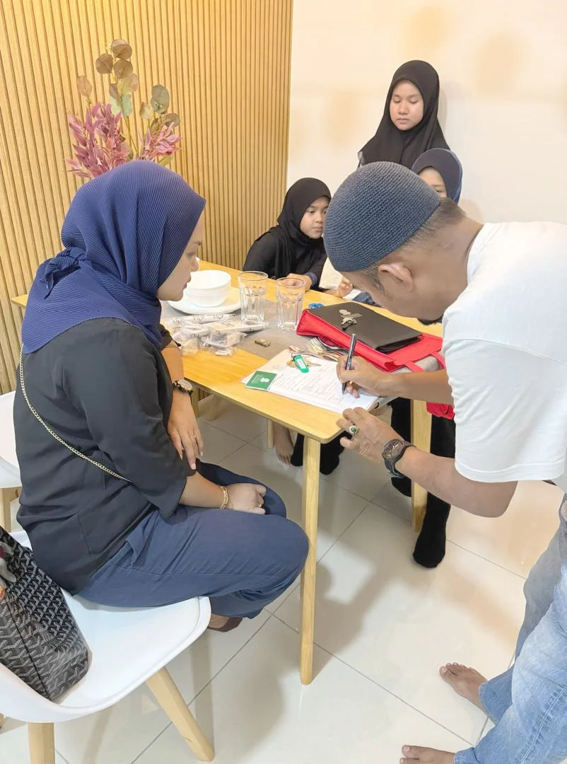 A group of women and a man are gathered around a table, engaging in a discussion or signing documents. The women are wearing black hijabs, and the man is wearing a gray cap and white shirt. The table has various items including glass cups, papers, a 