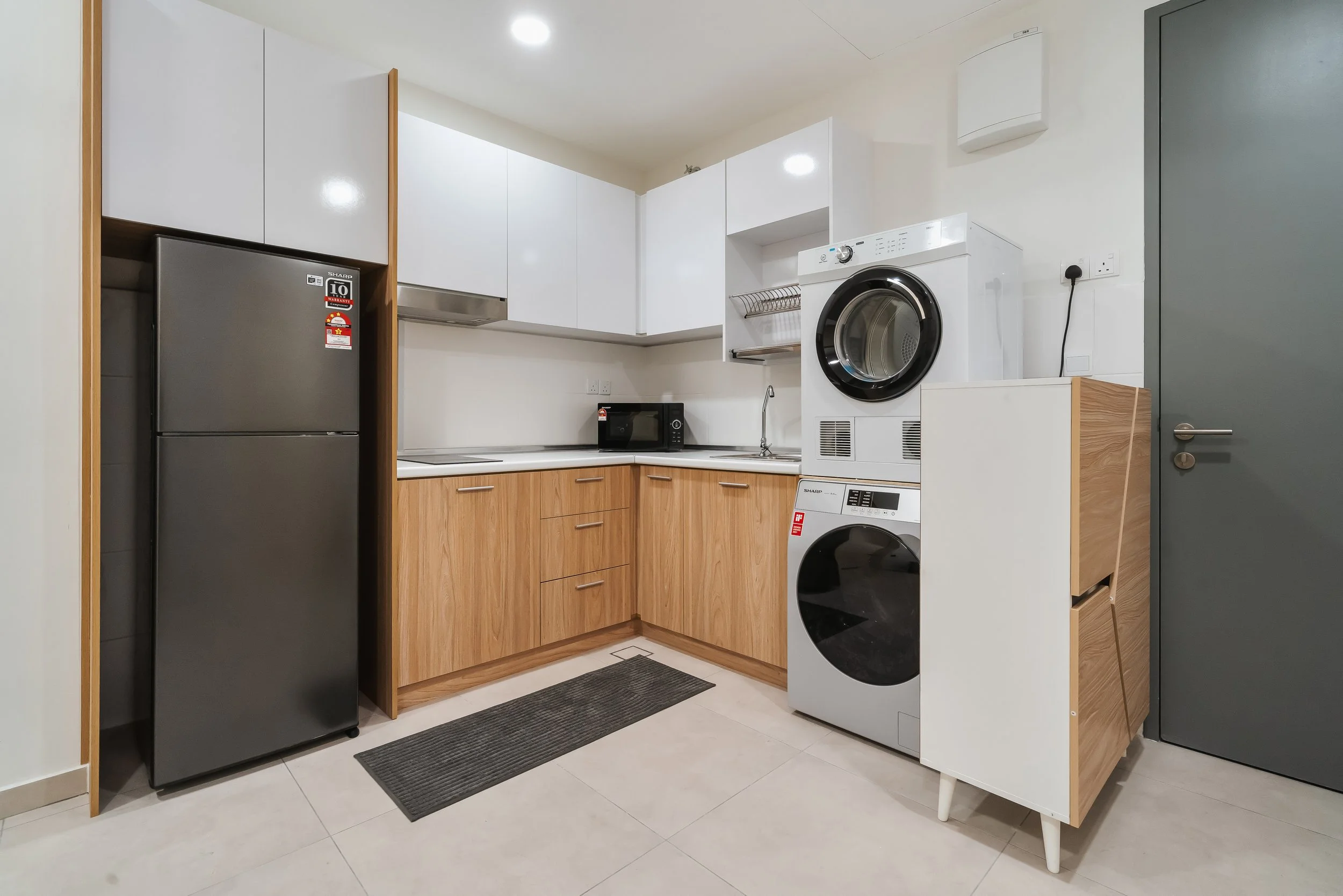 A compact kitchen with white and wooden cabinets, a black refrigerator, microwave, washing machine, and dryer, with gray tiled floors and a dark gray door.