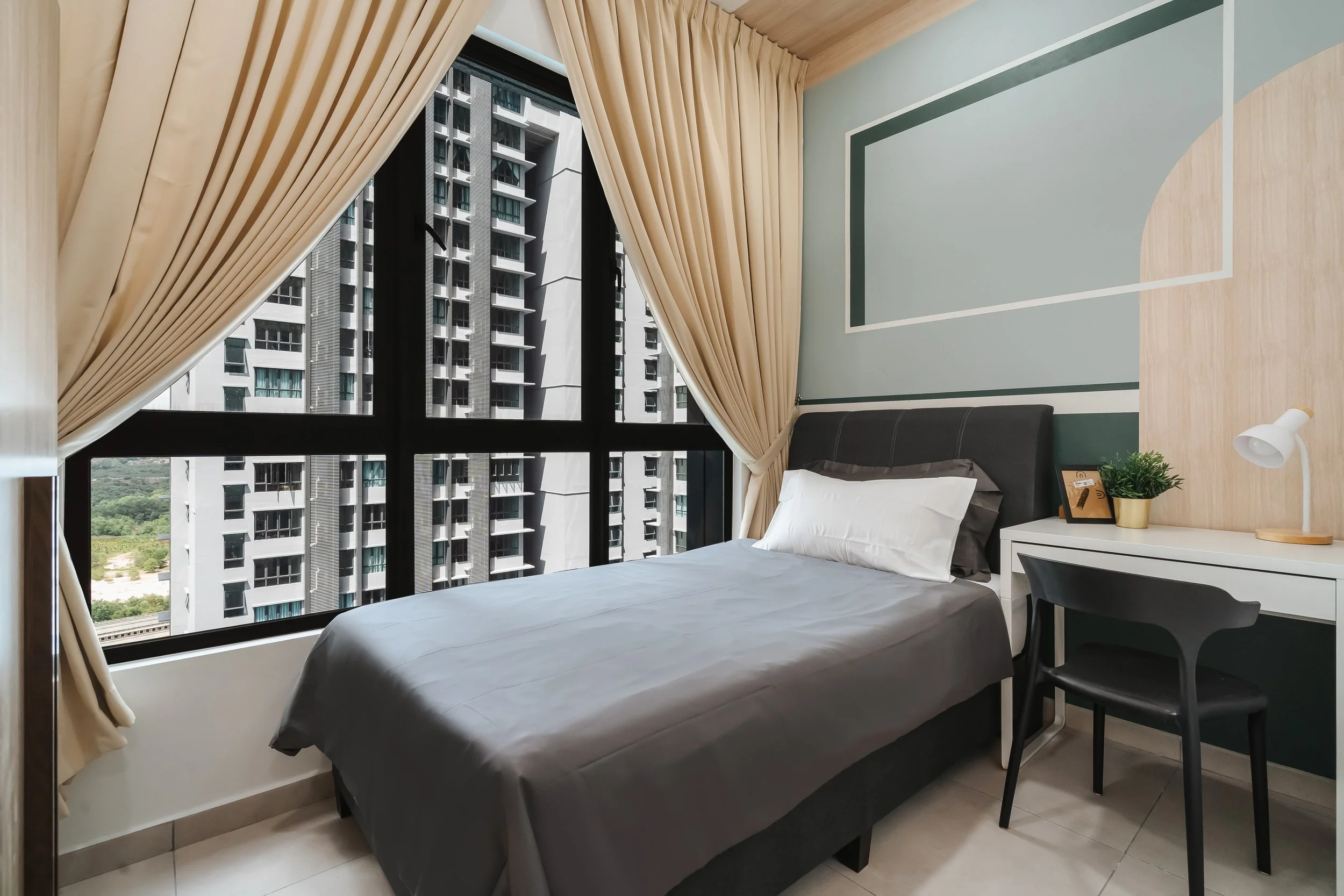 Interior view of a bedroom with a large window showing a high-rise building outside. The room has a bed with gray bedding, a white nightstand with a small plant, a book, and a white lamp. There is a black chair next to the nightstand and beige curtai