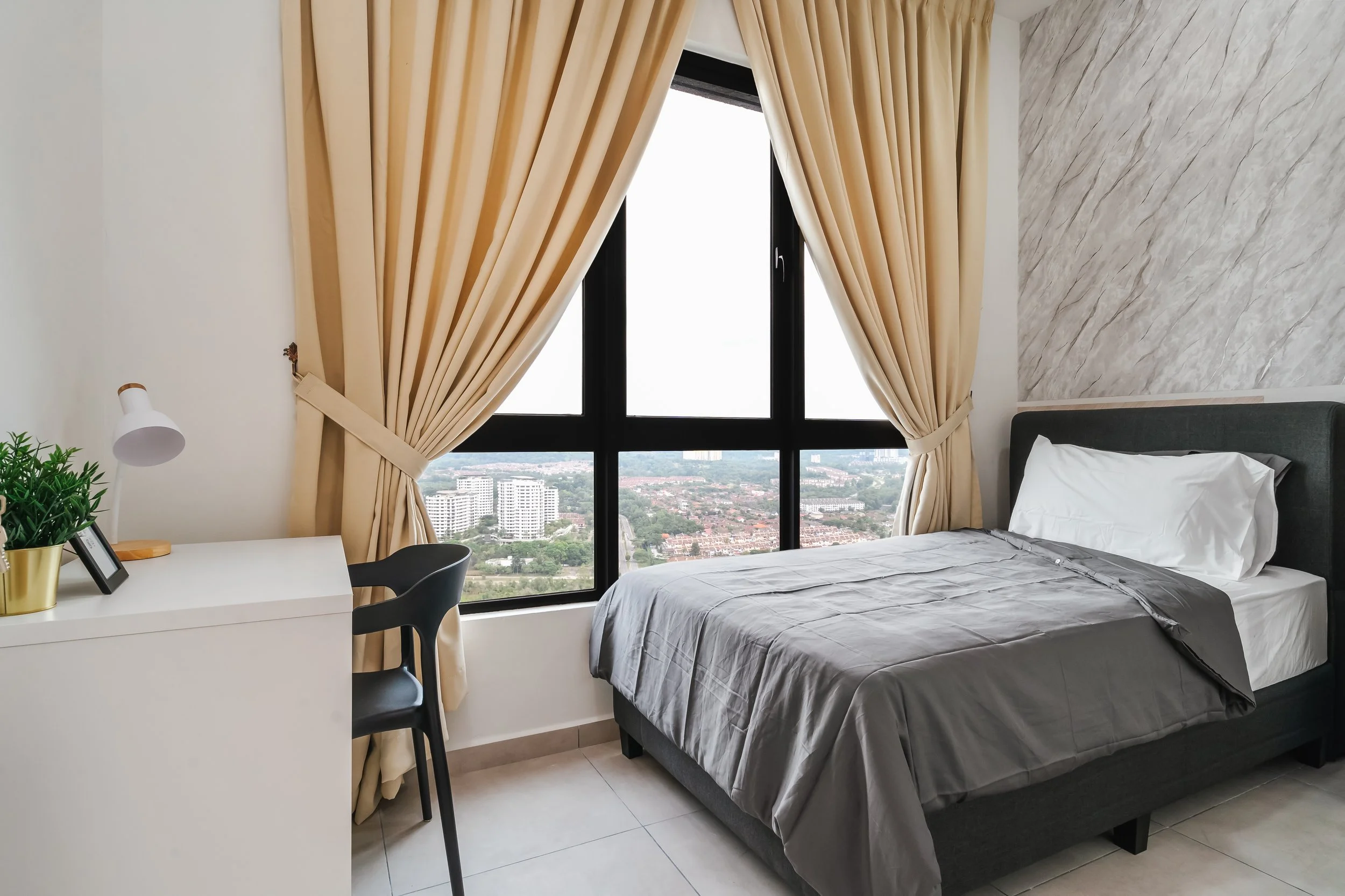 A modern bedroom with a large window, beige curtains, a white desk with a black chair, a potted plant, a watch, and a framed photo, with a bed and cityscape view outside.