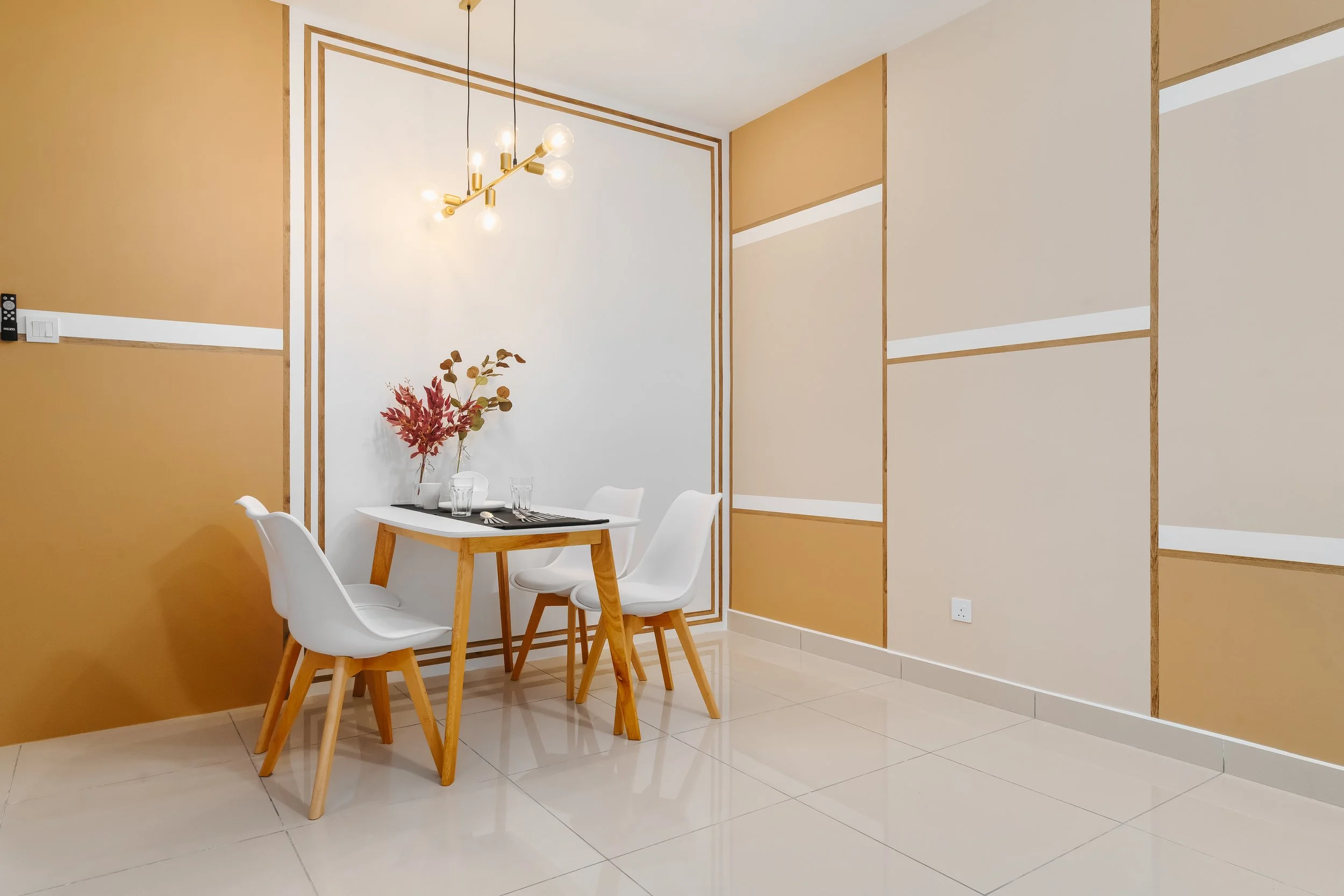 A modern dining area with a small wooden table set for two, surrounded by white chairs. The wall features a geometric design with beige and white panels outlined in wood framing. A contemporary chandelier with exposed bulbs hangs above the table, and a vase with autumnal flowers is on the table. The floor is tiled in a light color.