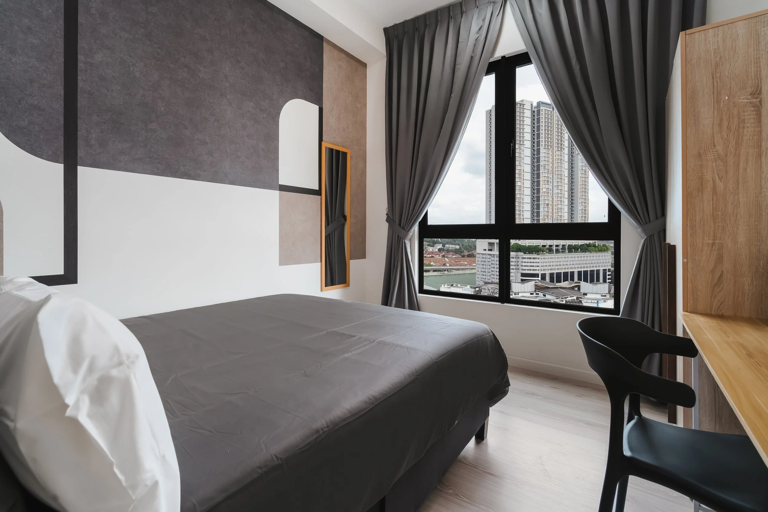 Modern hotel room with large window showing cityscape, gray curtains, a bed with gray bedding, a wooden desk, and a black chair.