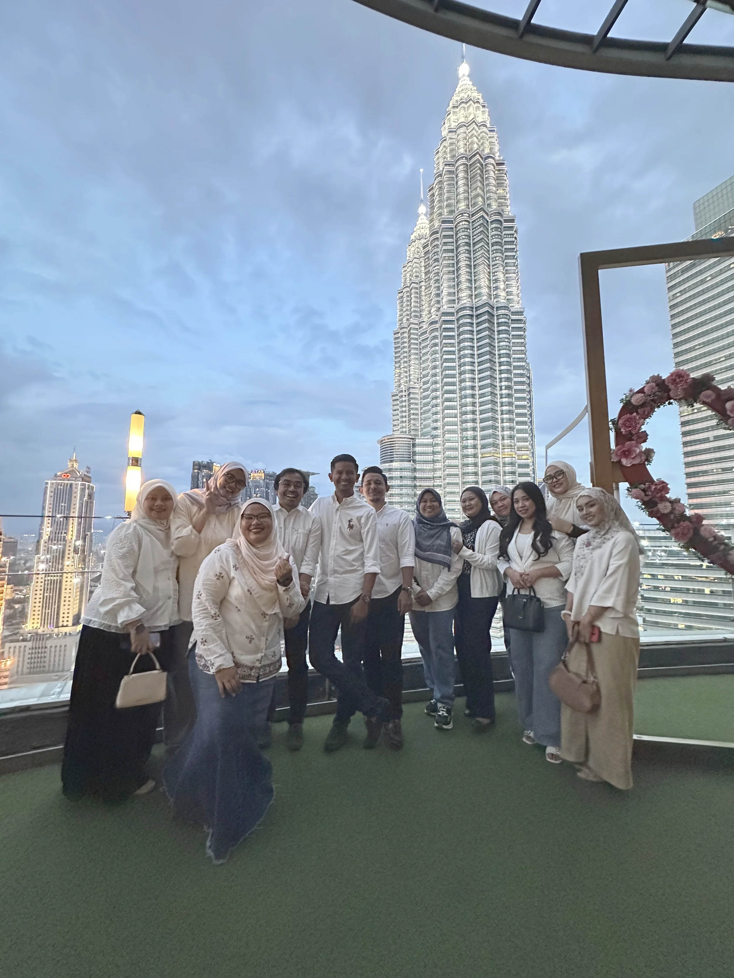 A group of people smiling and posing for a photo on a rooftop with a view of the Petronas Towers in Kuala Lumpur, Malaysia, during dusk.