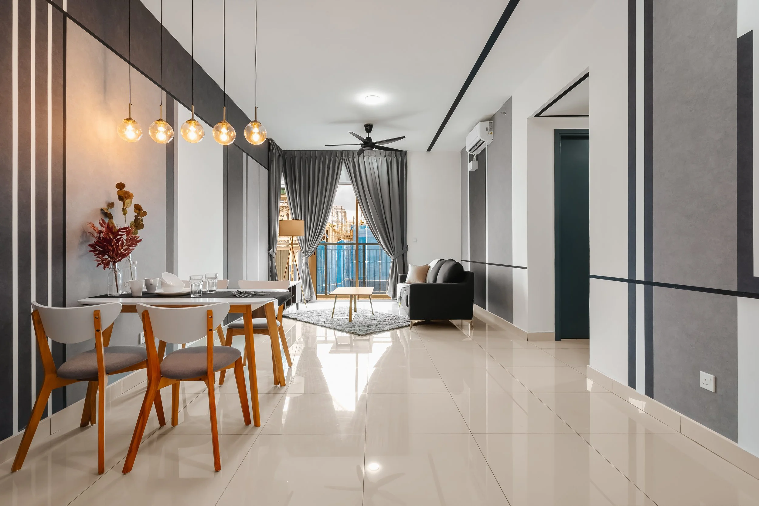 Modern living room with a dining area in the foreground, featuring a white dining table and chairs. The background includes a seating area with a black sofa, a round wooden coffee table, and large windows with gray curtains. The room has a ceiling fan and contemporary lighting fixtures with exposed bulbs, and the walls are decorated with gray and white vertical striped patterns.