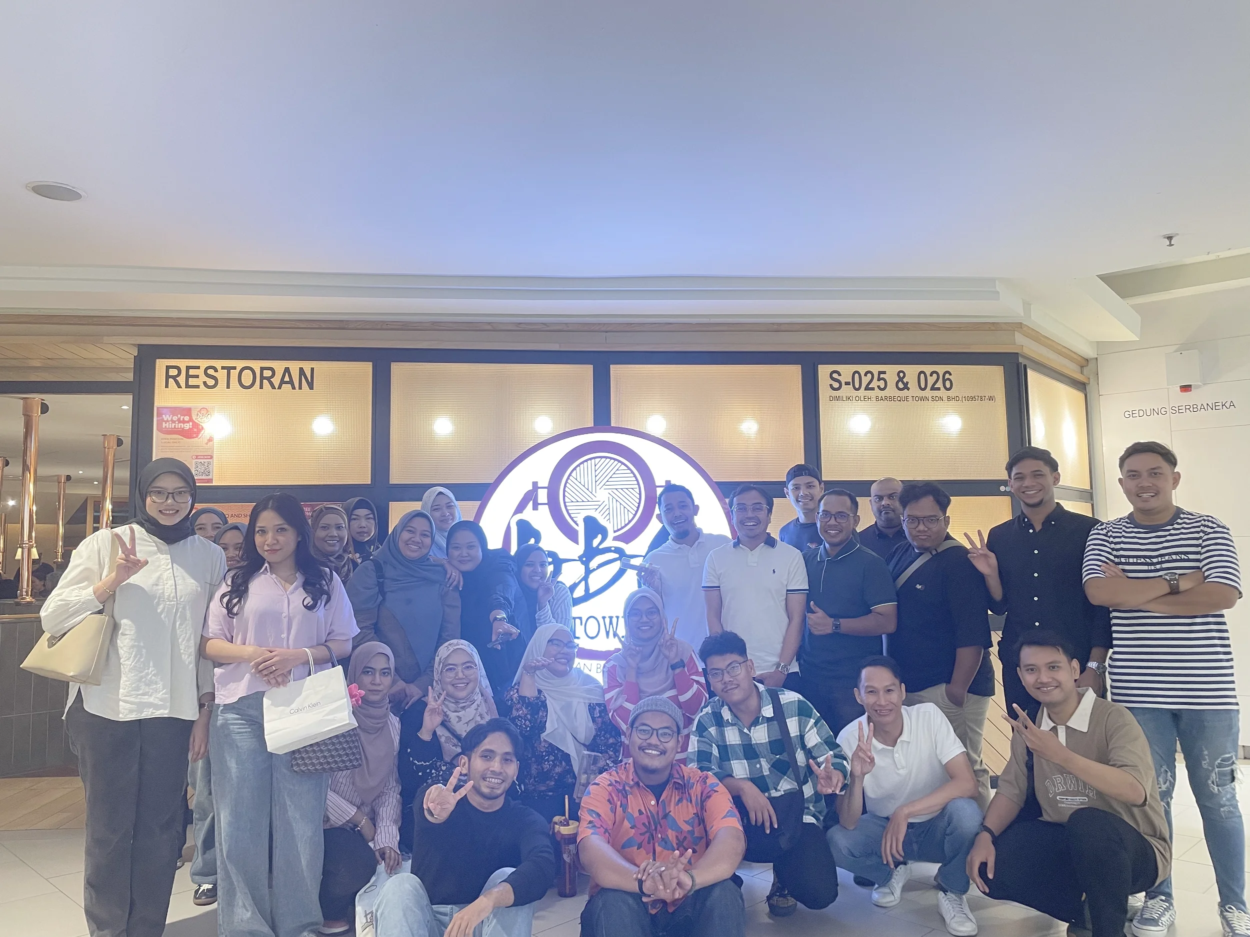 Group of diverse people posing and smiling at an indoor location, with a large illuminated sign behind them.