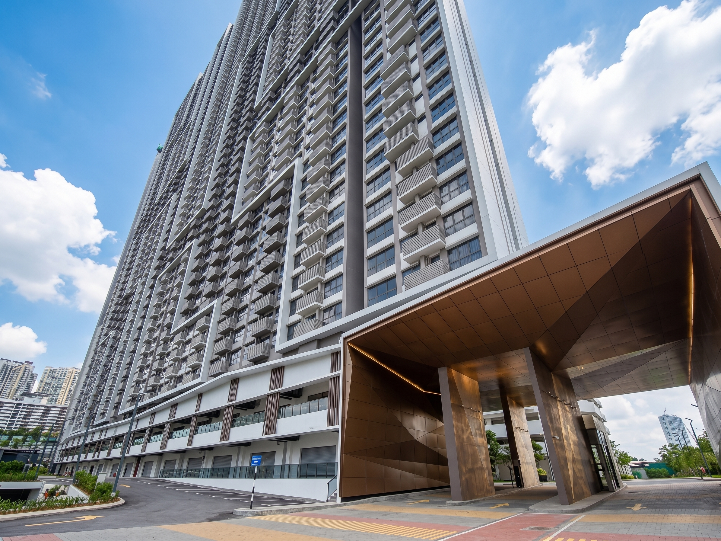 A modern high-rise building with white and gray exterior, multiple balconies, and large glass windows. The building features a distinctive overhang at the entrance with a gold-colored underside and integrated lighting. The surroundings include a parking lot and a clear sky with few clouds.