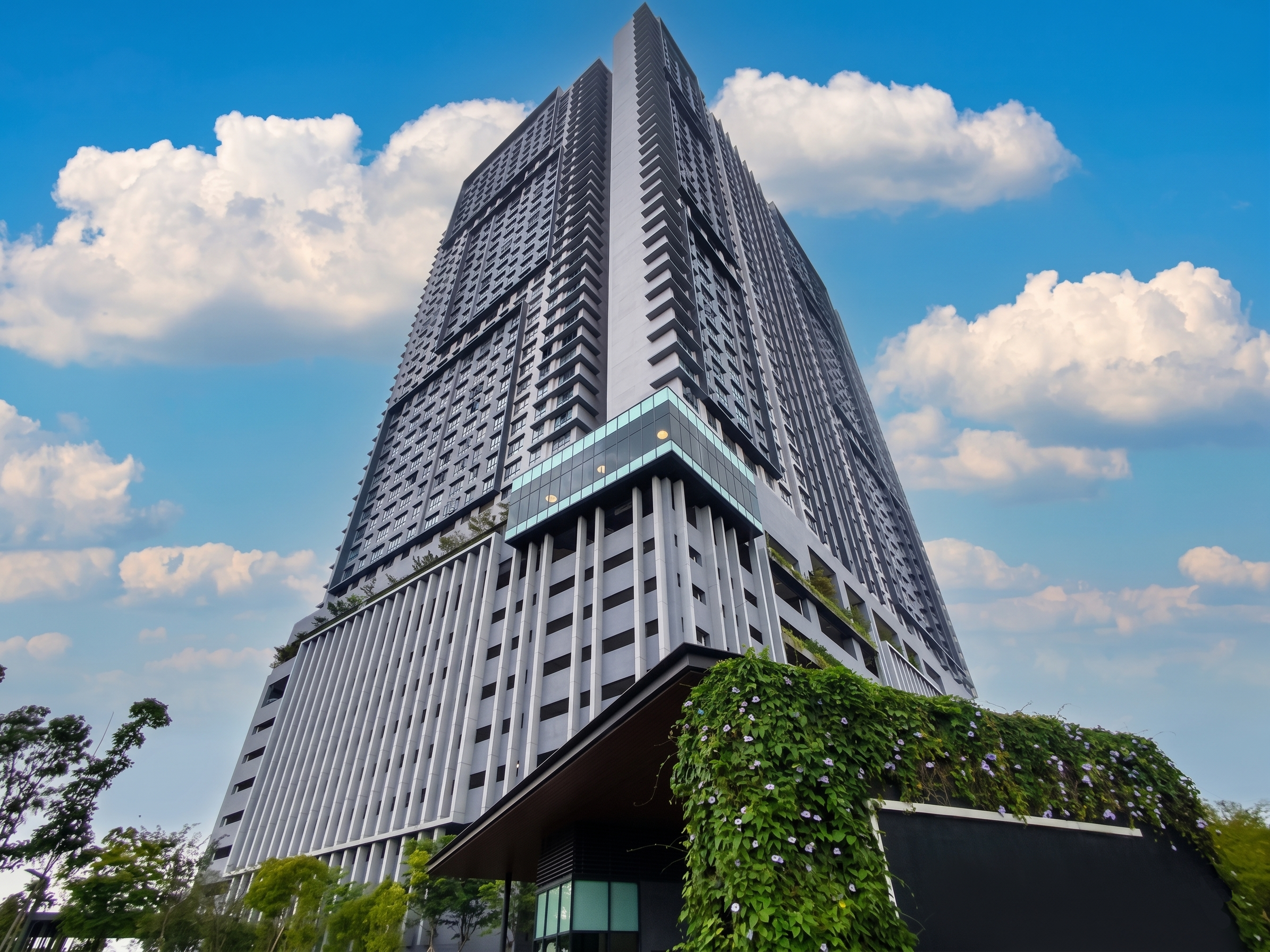 A tall modern skyscraper with a black and gray facade, glass balcony, and vertical white slats at the base, under a partly cloudy blue sky.