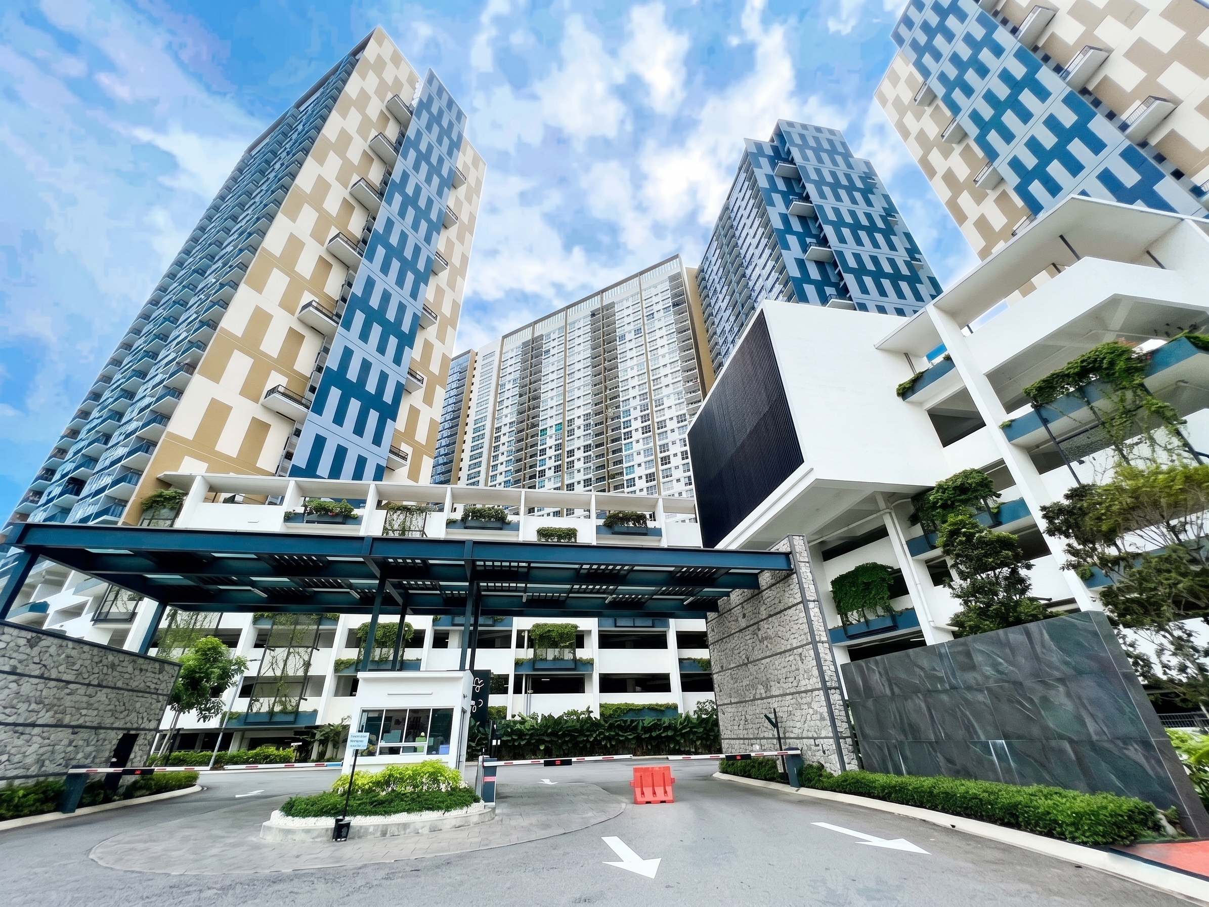 Modern high-rise residential buildings with glass balconies, set against a blue sky with clouds, featuring a parking area with a barrier and landscaped greenery at the entrance.