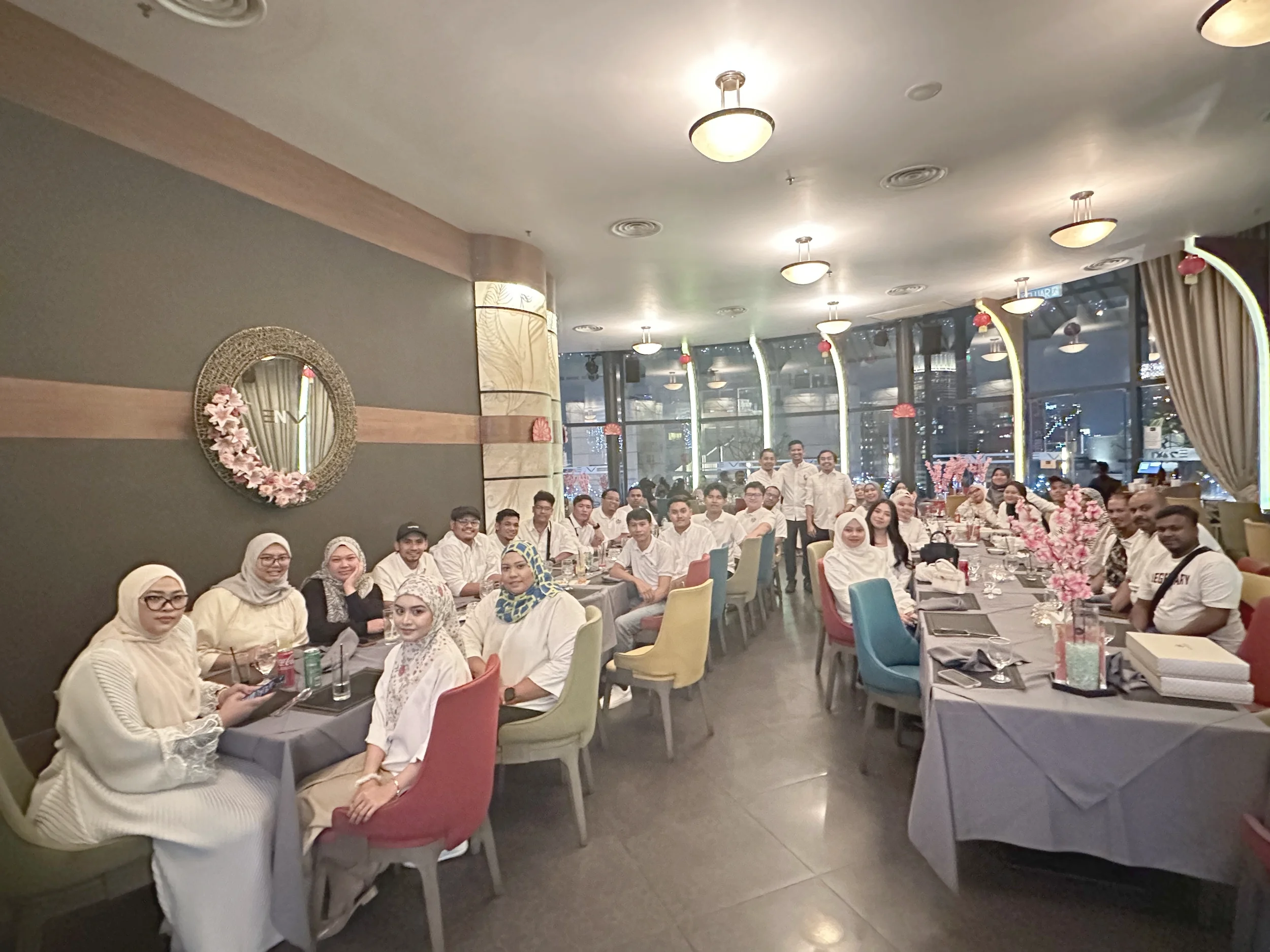 Group of people gathered in a restaurant, seated at tables with pink cherry blossom centerpieces, with a large glass window showing a city scene outside, and several ceiling lights illuminating the room.