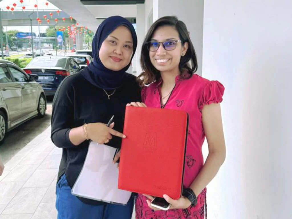 Two women standing outdoors, one wearing a black hijab and blouse, the other wearing glasses and a pink dress, holding a red document folder.