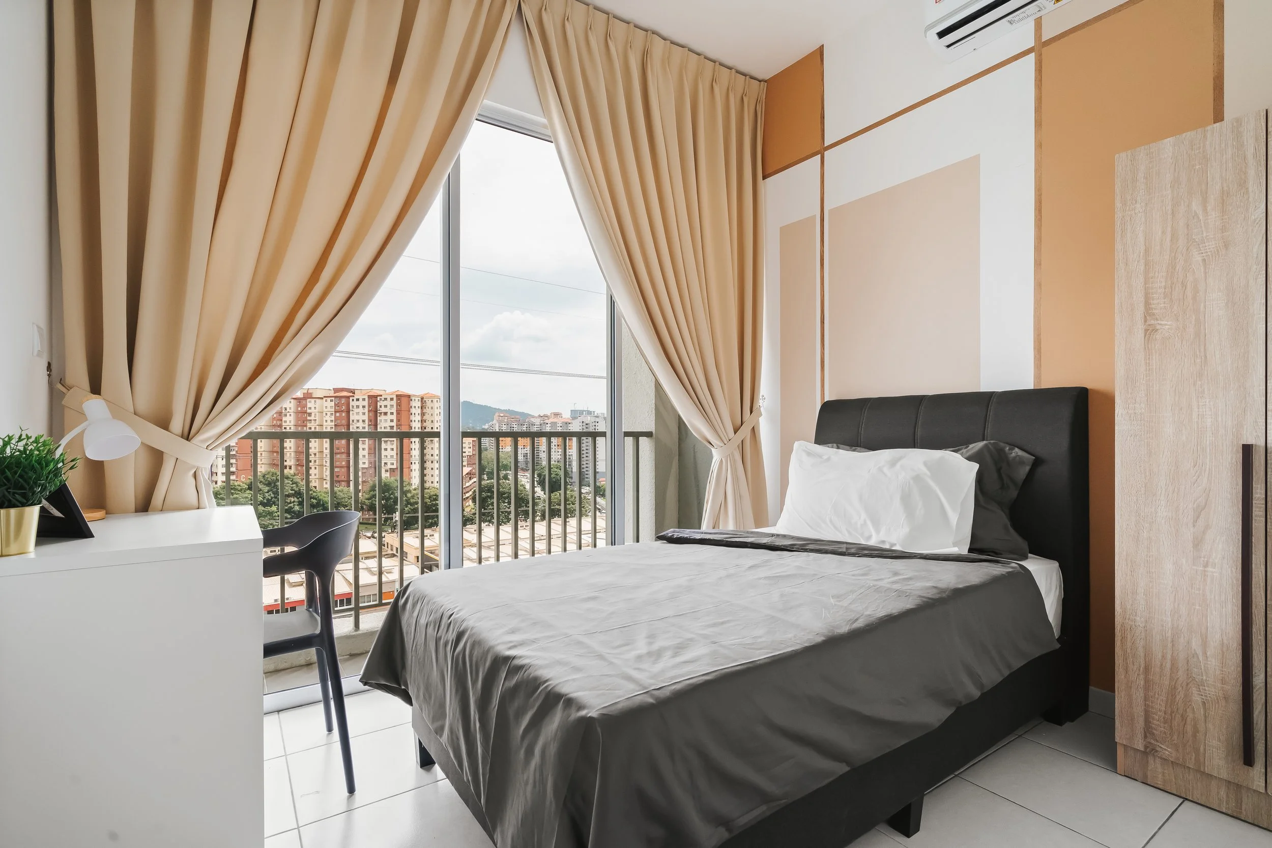 A bedroom with a bed, beige curtains, a small desk with a plant and lamp, and a large window overlooking a cityscape with high-rise buildings and mountains in the background.