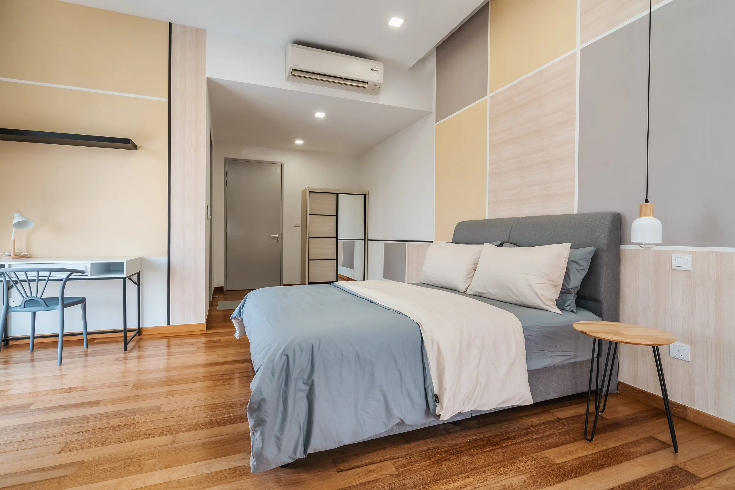 Modern bedroom with wooden floors, a bed with gray bedding, a gray upholstered headboard, beige and gray accent walls, a small wooden side table, and a white and wooden lamp hanging from the ceiling. There is a small desk with a chair and a shelf on the left side, and a metal framed sliding door closet and air conditioning unit on the back wall.