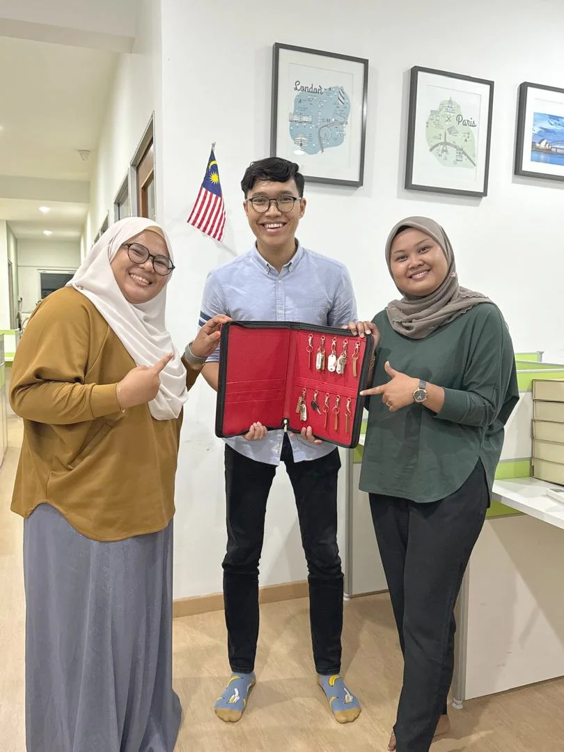 Three people smiling, two women and one man, standing indoors with a Malaysian flag and framed pictures of Paris and London on the wall behind them. The man is holding a red key case with multiple keys, and all three are pointing at it.