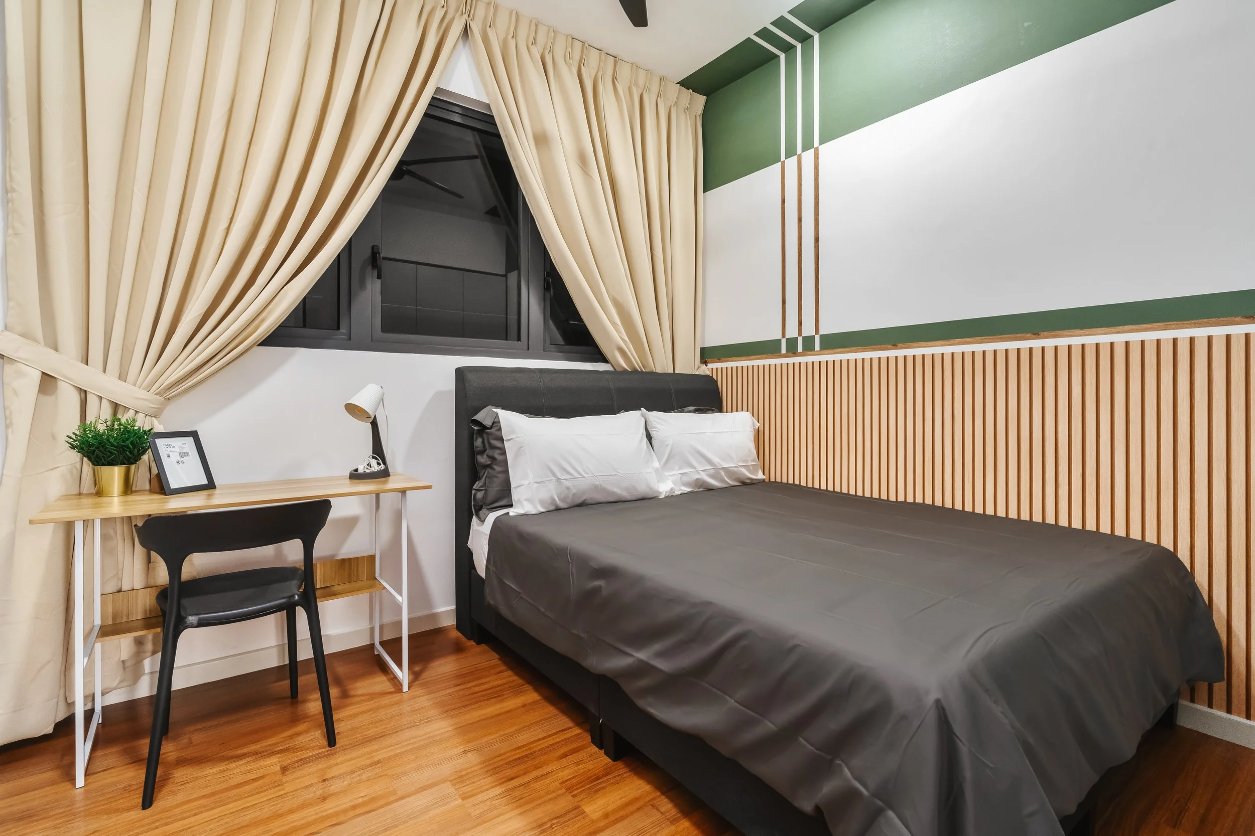 A bedroom with beige curtains, a window, a bed with dark gray bedding, a wooden desk with a black chair, a potted plant, a picture frame, and a wall painted green and white with wooden paneling.