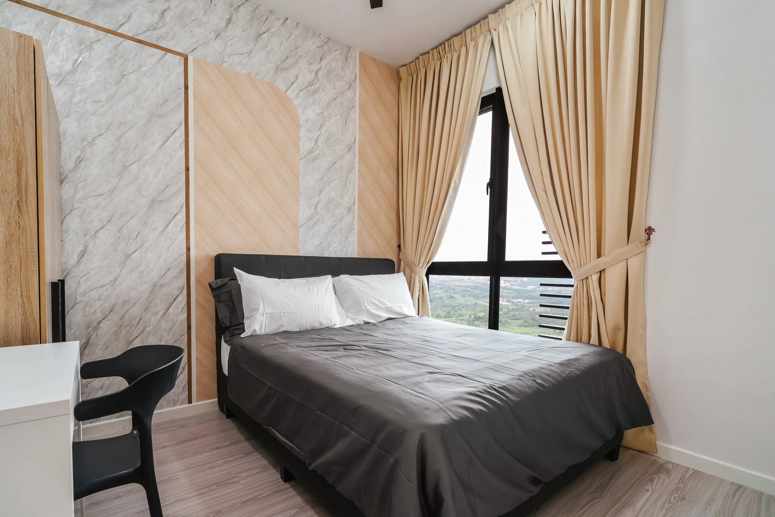A modern bedroom with a large window covered by beige curtains, a black bed with white pillows and gray bedsheet, a white small table, and a black chair, with a view of green landscape outside.