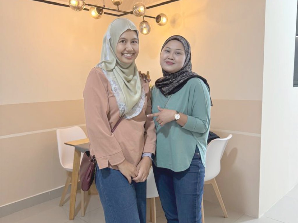 Two women standing indoors in a cozy, well-lit space with modern decor, both wearing hijabs and casual outfits, smiling at the camera.