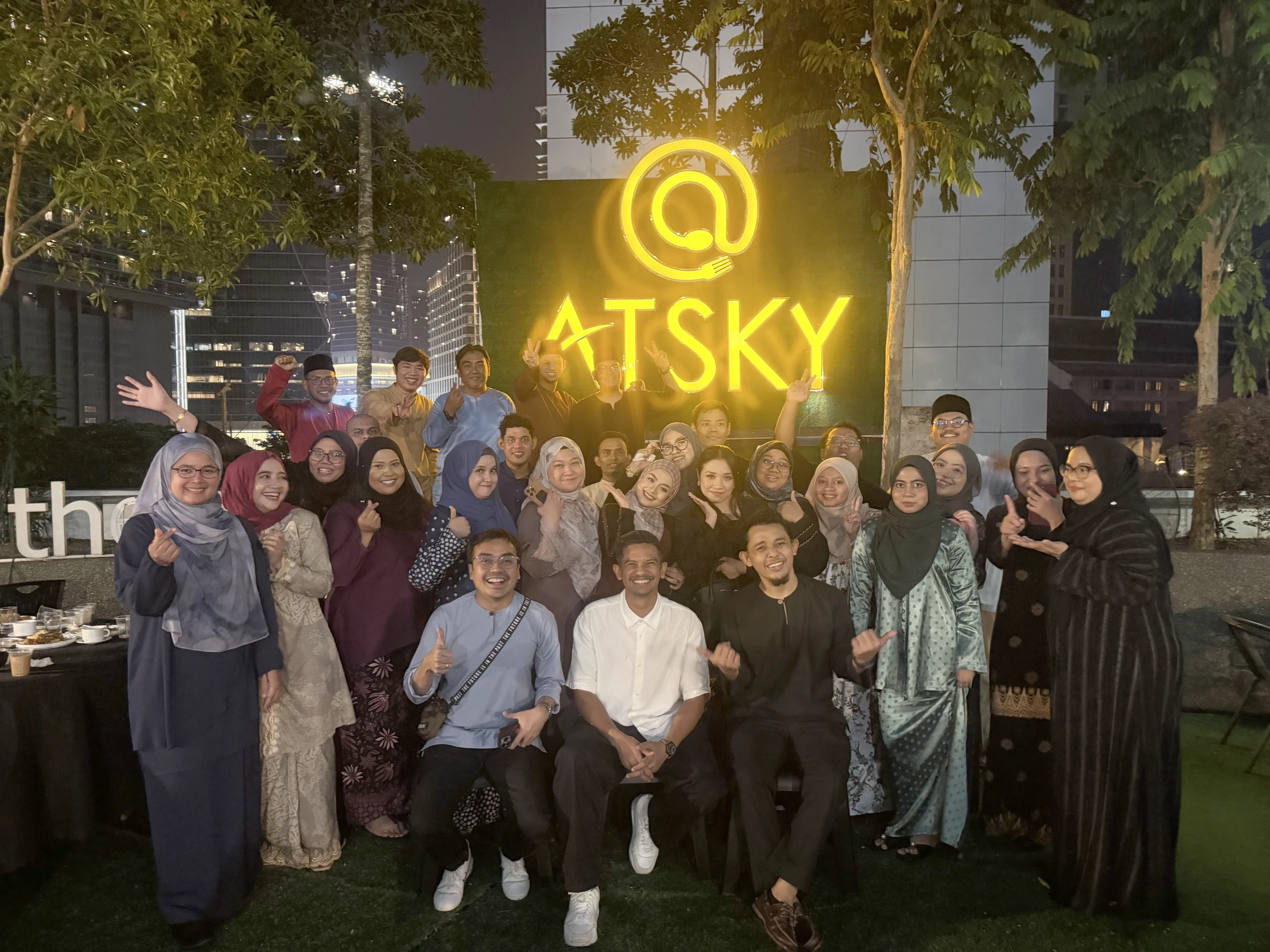 Group of people gathered outdoors at night, smiling and making gestures, in front of a large illuminated yellow sign with a stylized A and the word "ATS Ky" and trees in the background.