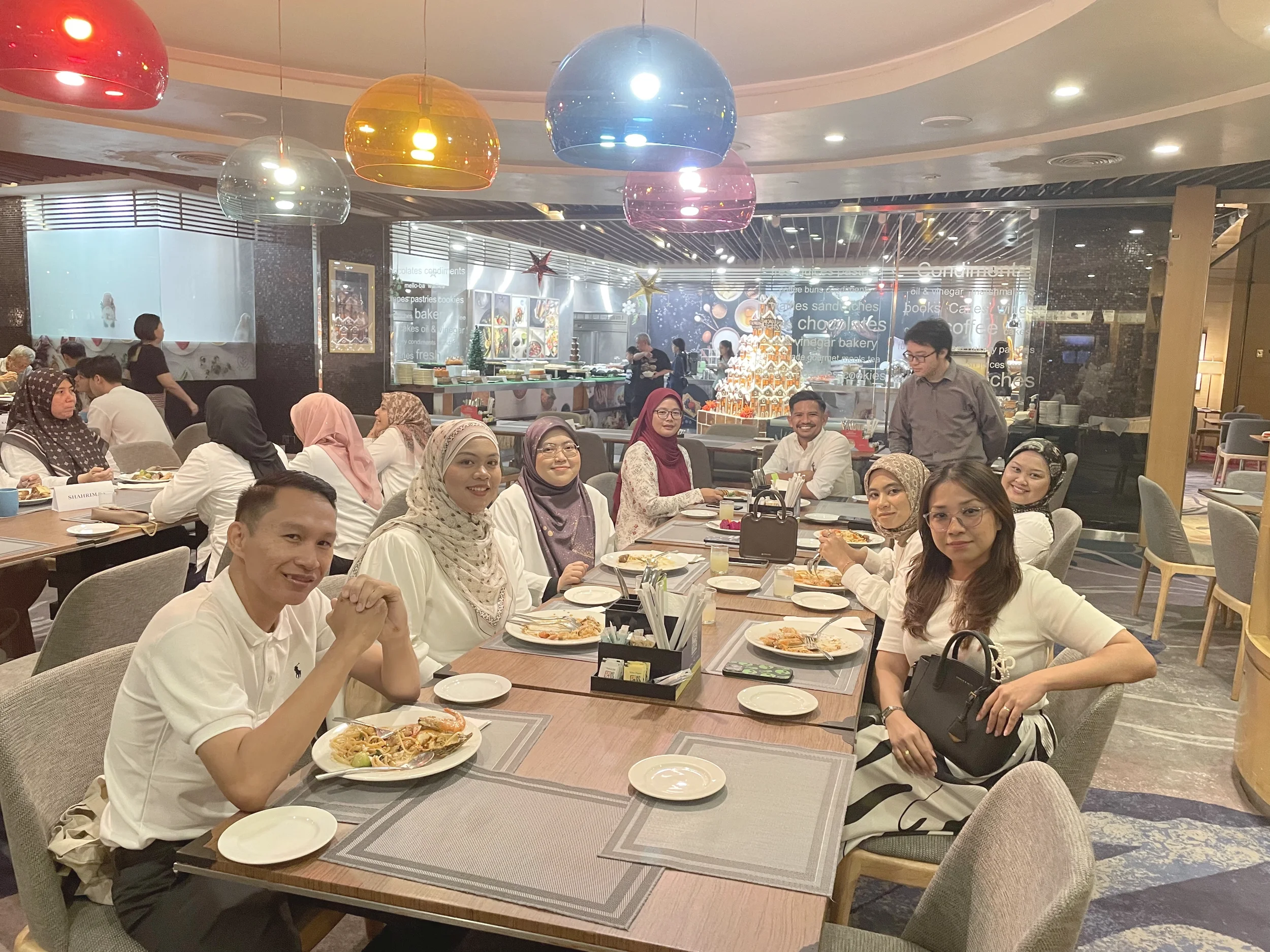 People sitting at tables in a restaurant, enjoying a meal with various dishes. Decor includes colorful hanging lights, a festive gingerbread house display, and wall art. The group appears to be celebrating or socializing.
