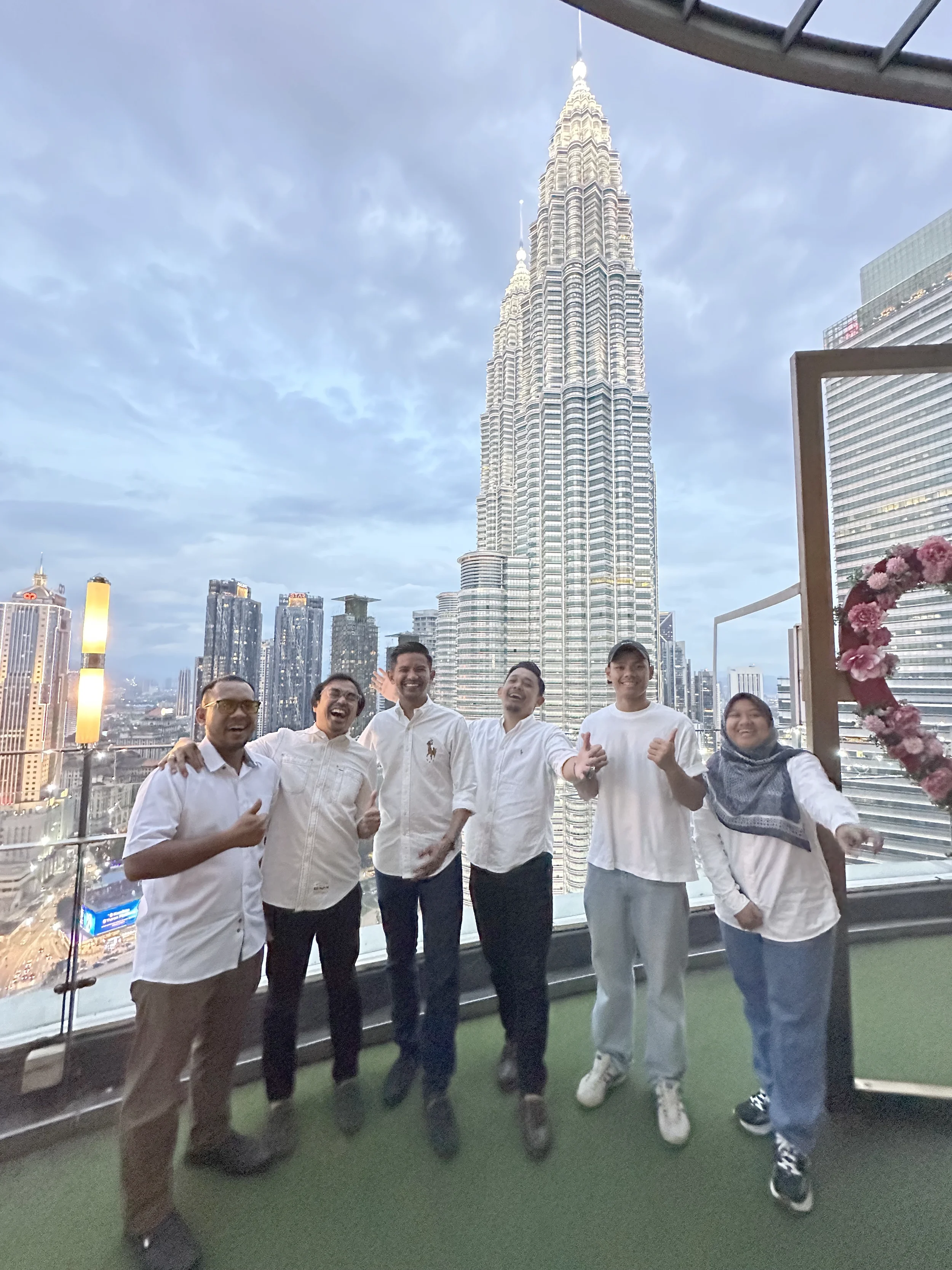 Group of six people smiling and posing for a photo on a balcony with the city skyline, including the Petronas Towers, in the background.