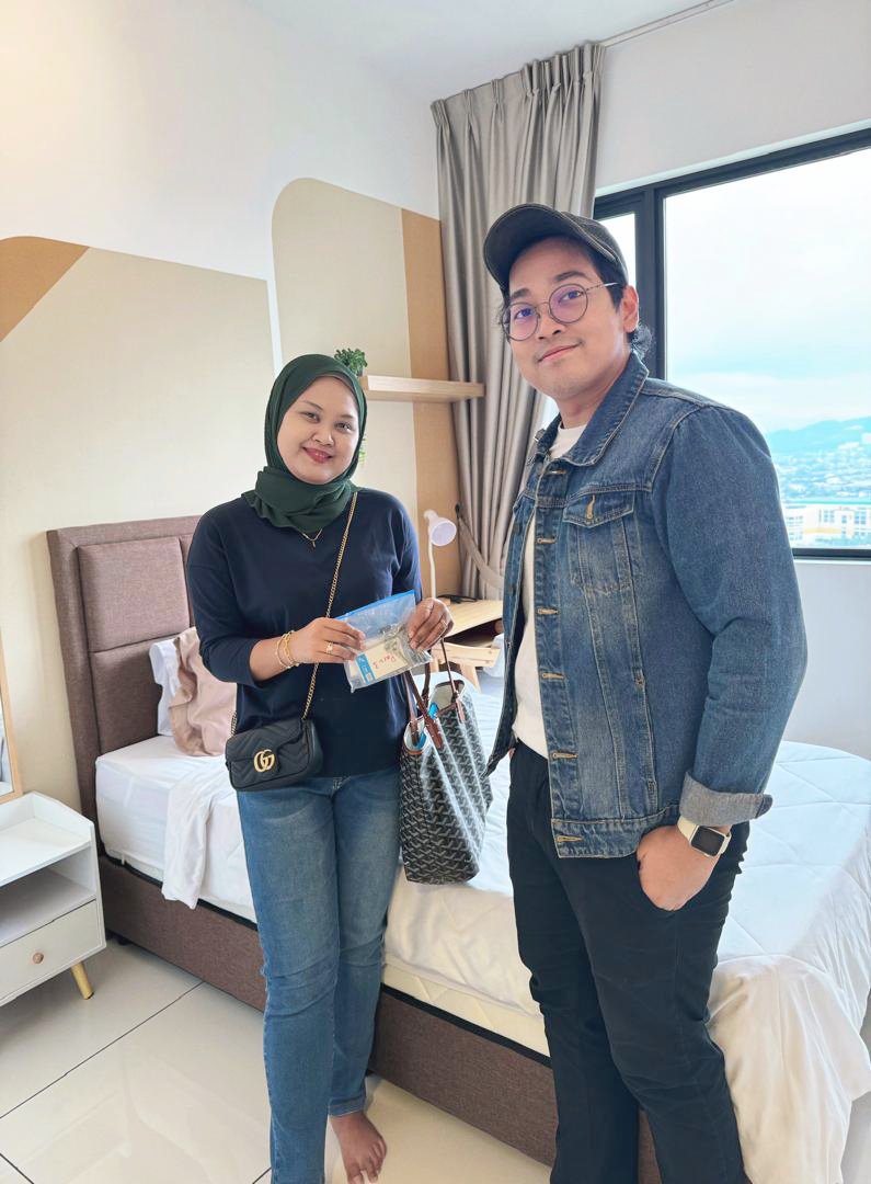 A woman and a man stand in a bedroom, smiling at the camera. The woman wears a dark green headscarf, navy top, and jeans, holding a plastic bag. The man wears a denim jacket, white shirt, and black pants, with a watch on his wrist. The bedroom has a 